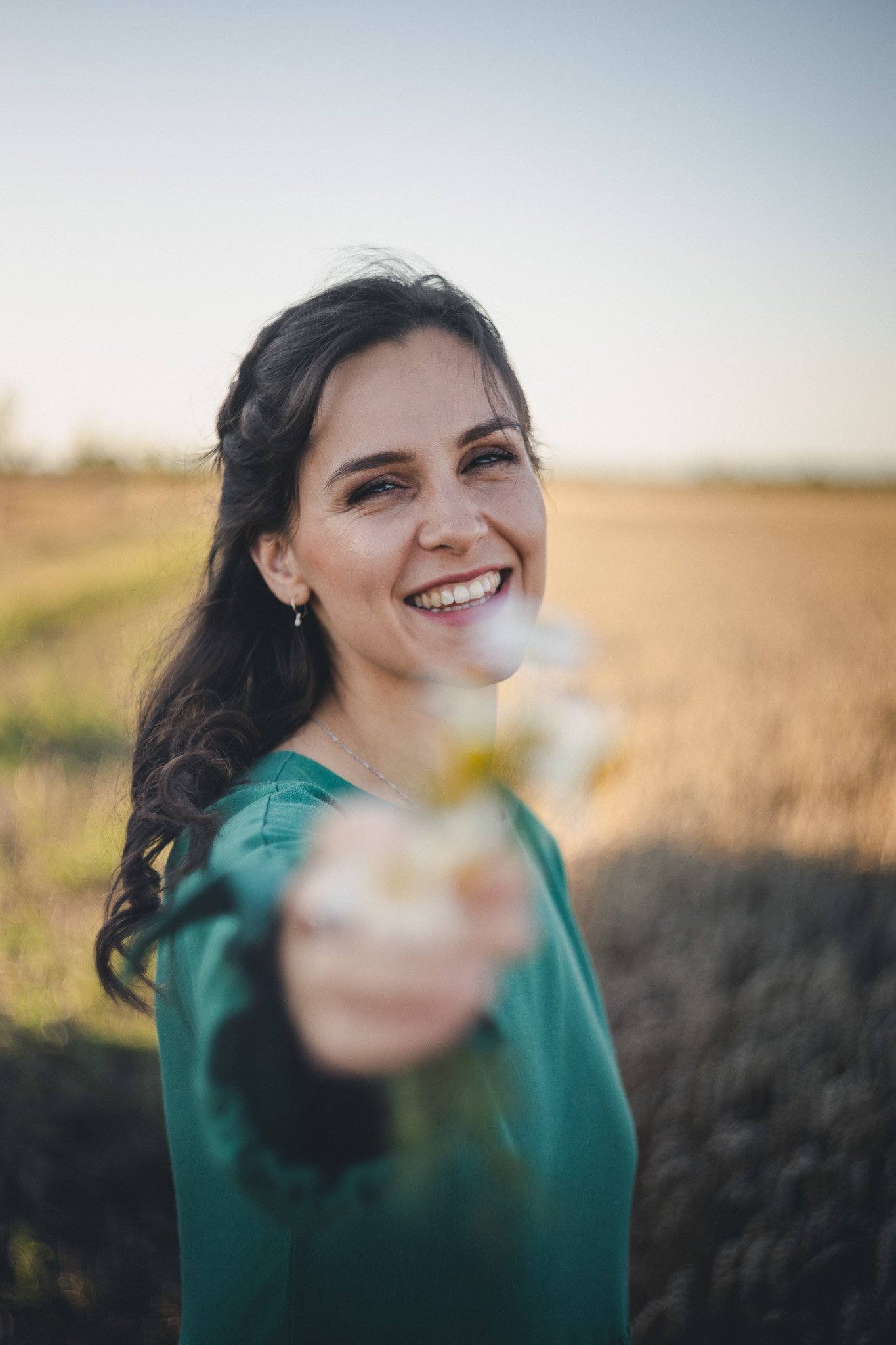 A smiling woman with dark hair, wearing a teal top, extends her hand with a bouquet, standing outdoors in a field during sunset.