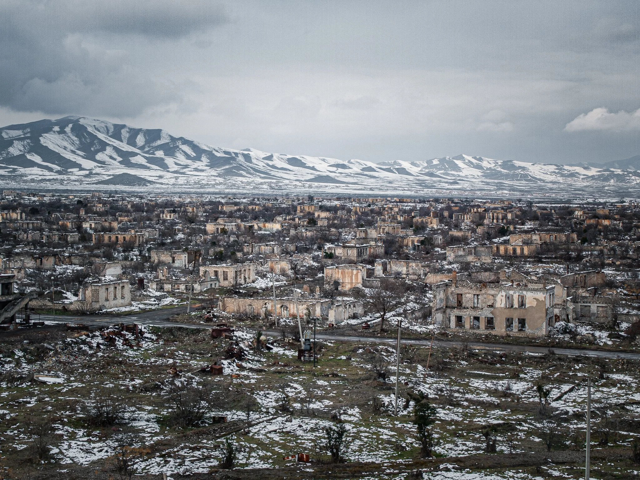 Desolate landscape with abandoned, damaged buildings and snow-covered ground, mountain range in the background under a cloudy sky.