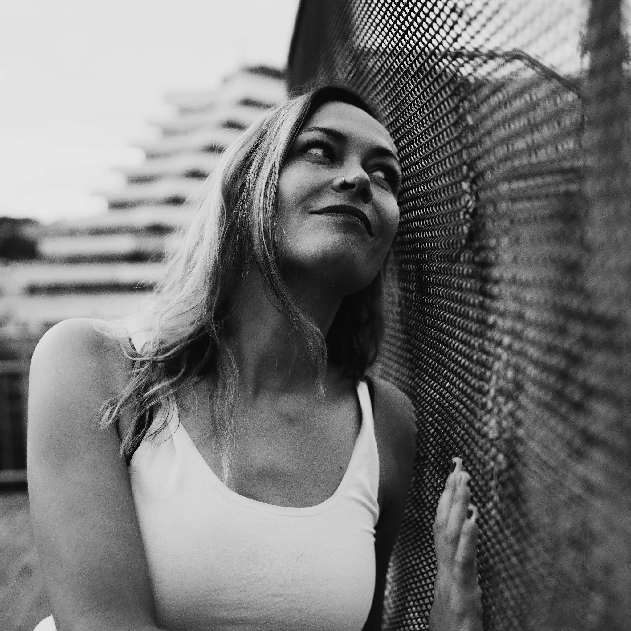 A woman with shoulder-length hair wearing a sleeveless top, leaning her head against a chain-link fence, smiling softly, in black and white.