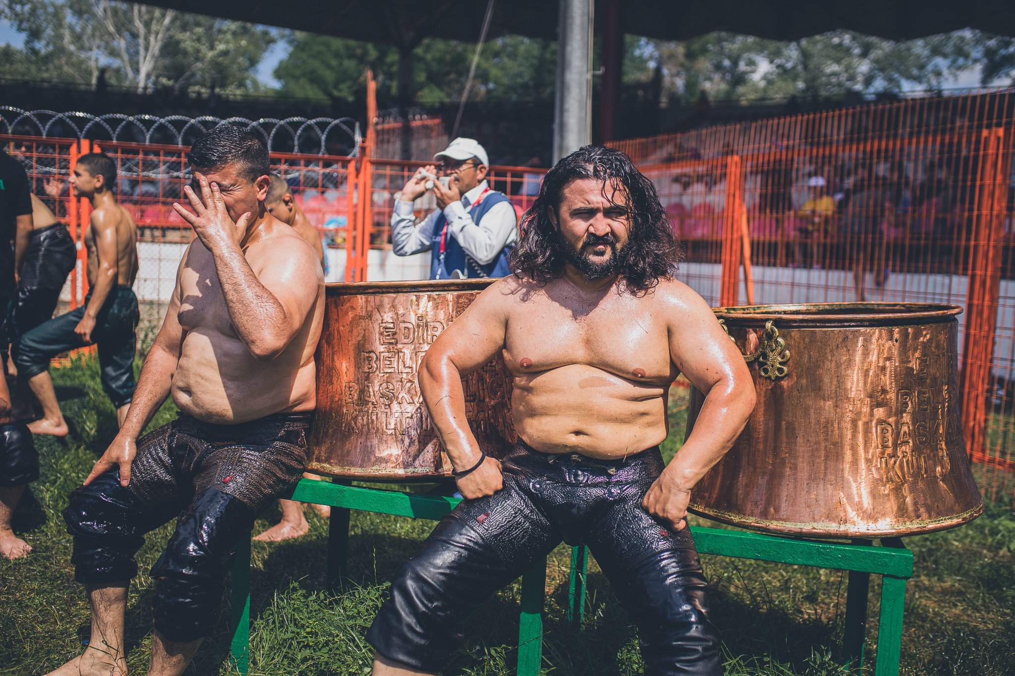 Two shirtless men with long hair and leather pants sitting on a green bench, with copper washbasins behind them, in an outdoor setting surrounded by a fence and other people.