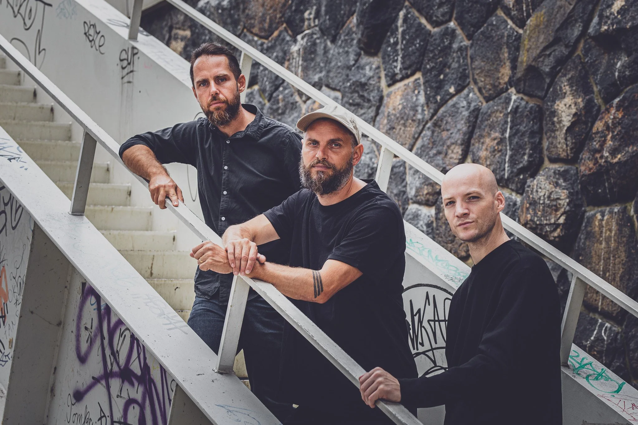 Three men with beards standing on graffiti-covered stairs outdoors, with a dark stone wall in the background.
