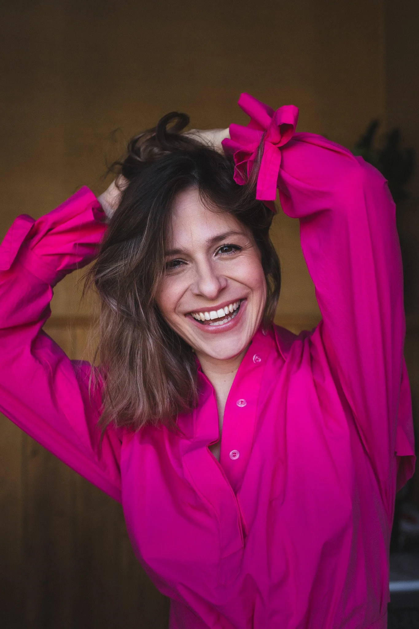 A woman with shoulder-length brown hair smiling and wearing a bright pink shirt, with her arms raised and hands behind her head.