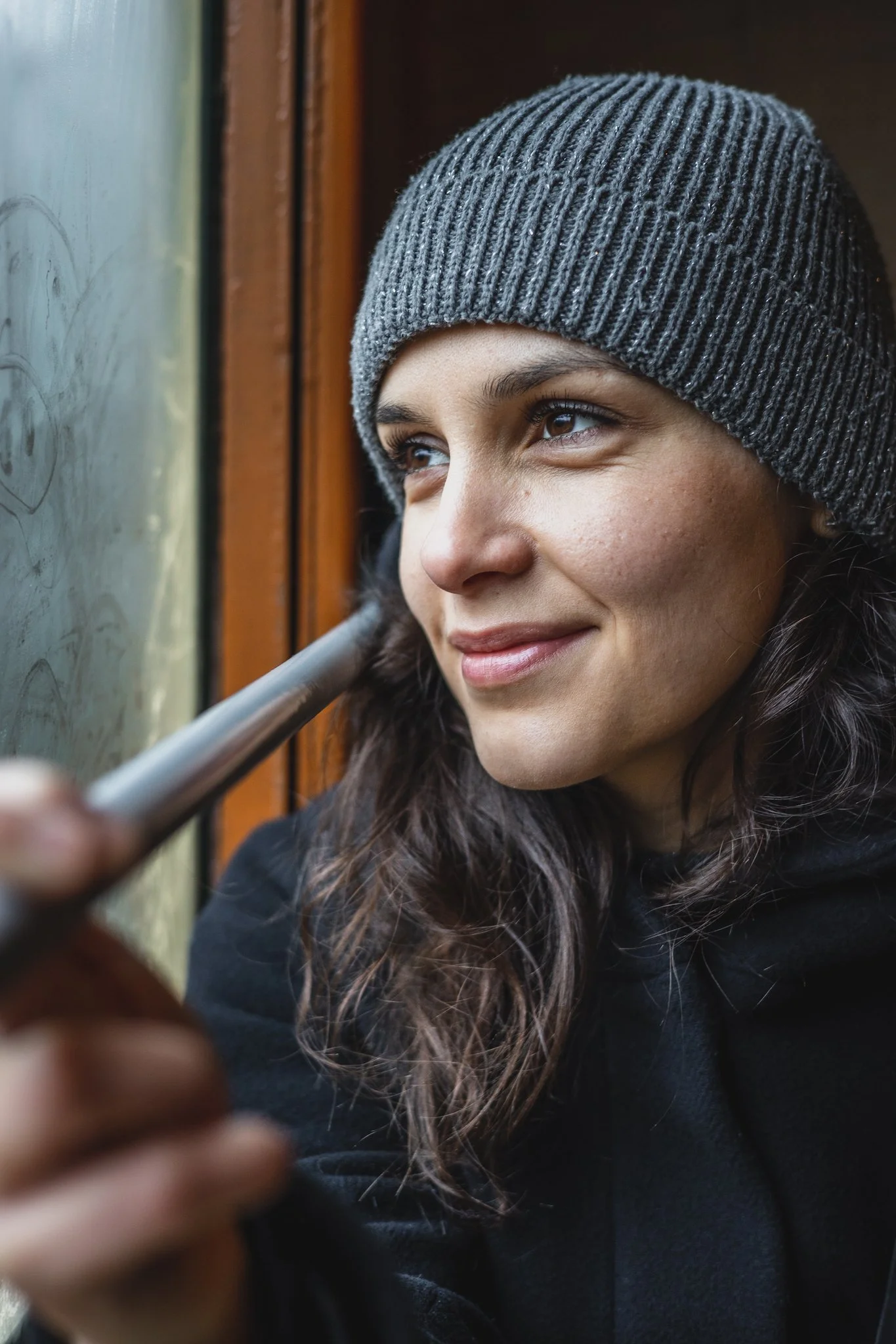 A young woman in a knit gray beanie smiling while looking out a window, holding a phone.