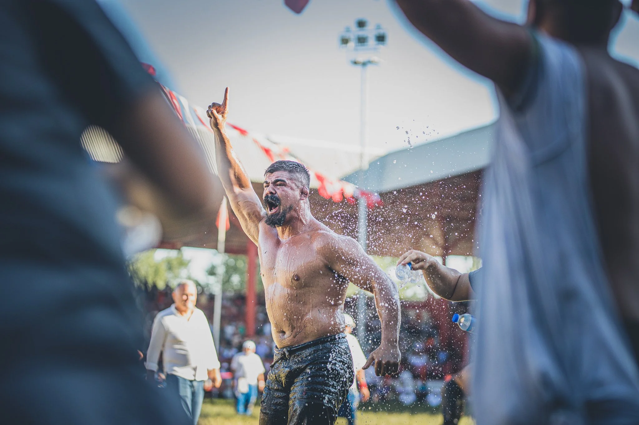 A shirtless man with a beard and short hair shouting and raising his right hand in the air during an outdoor event, with water splashing around him and people in the background.