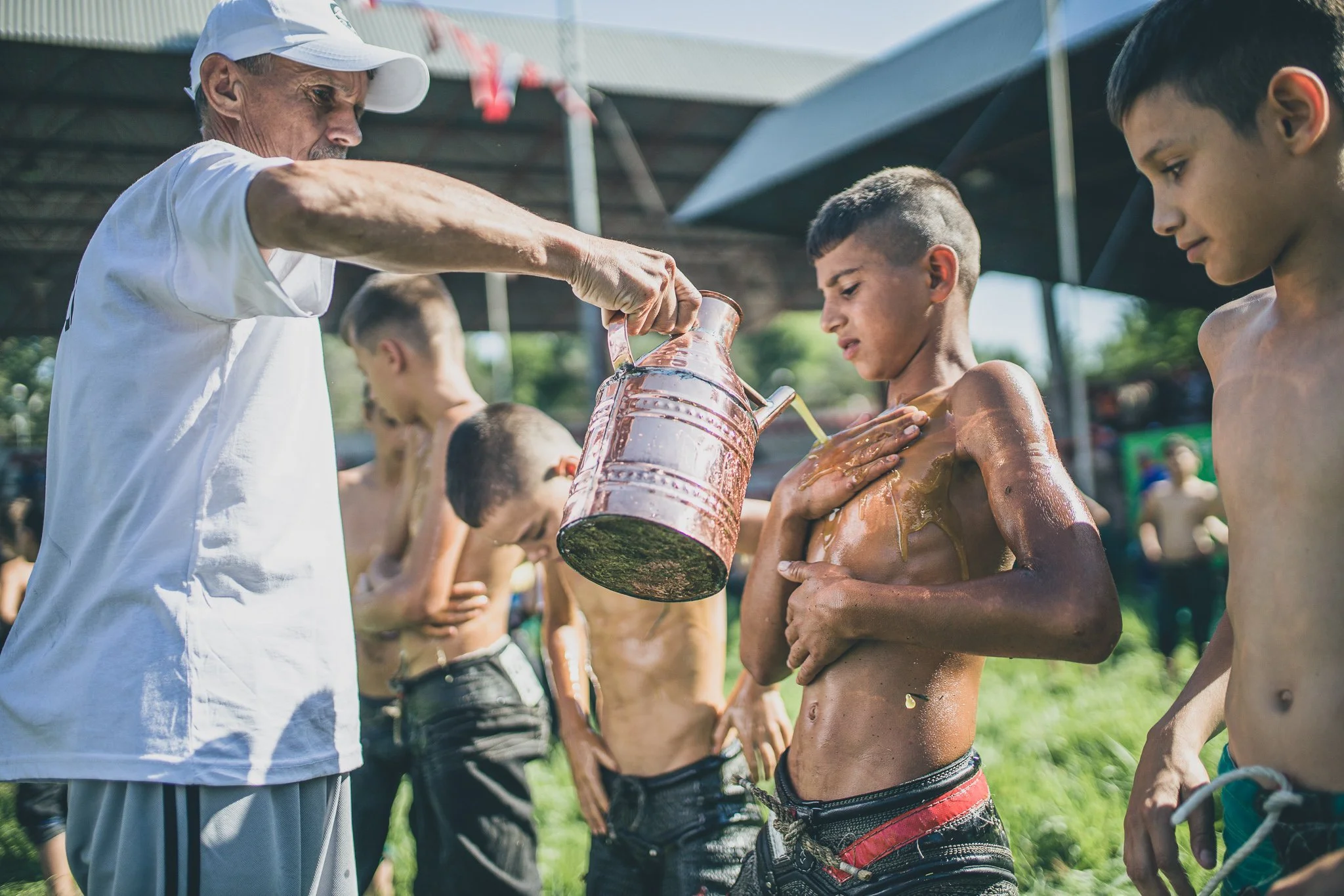 An older man pouring water over a group of shirtless boys standing in line under a shaded outdoor area.