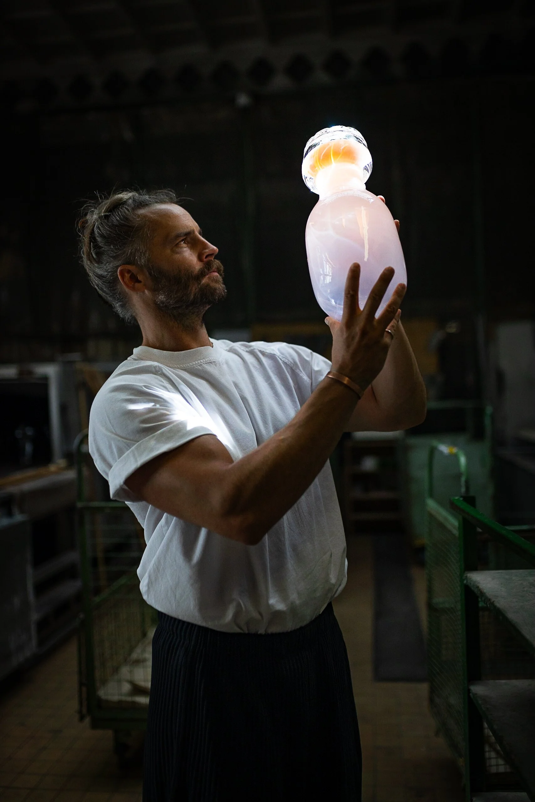 A man with a beard and long hair in a bun, wearing a white t-shirt, holding and examining a glowing, translucent glass sculpture in a dimly lit workshop.
