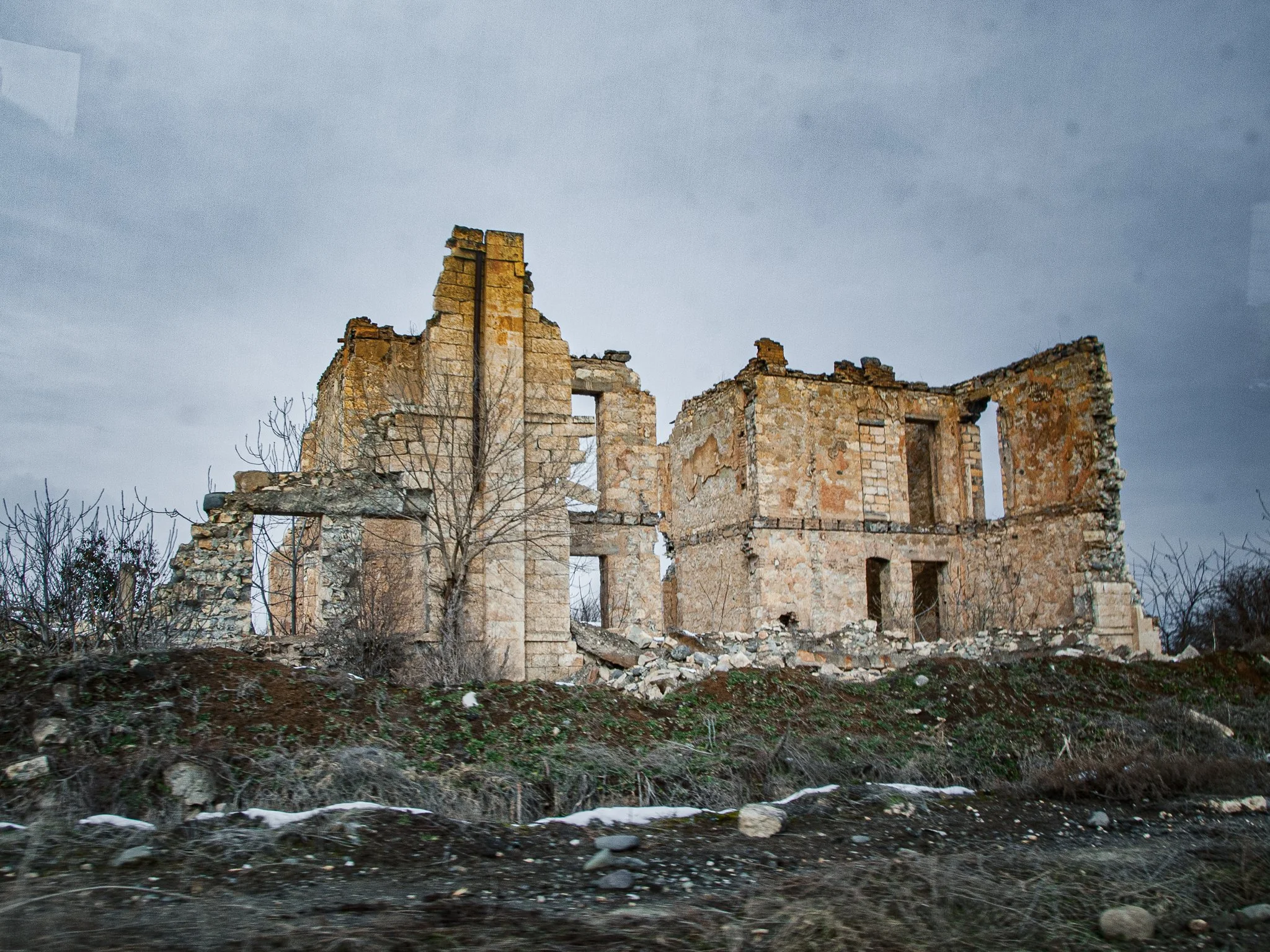 Ruined stone house with missing roof and walls, set on a small hill with sparse vegetation and leafless trees, under a cloudy sky.
