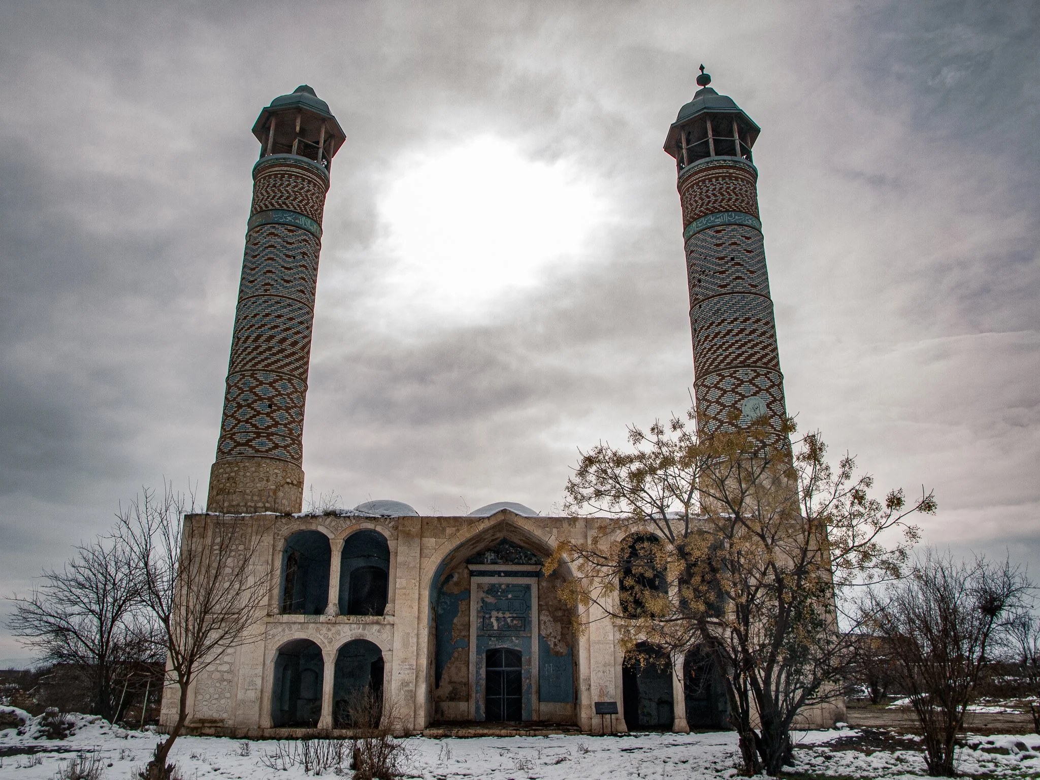 A historic building with two tall, intricately decorated minarets under a cloudy sky, with snow on the ground and leafless trees in the foreground.