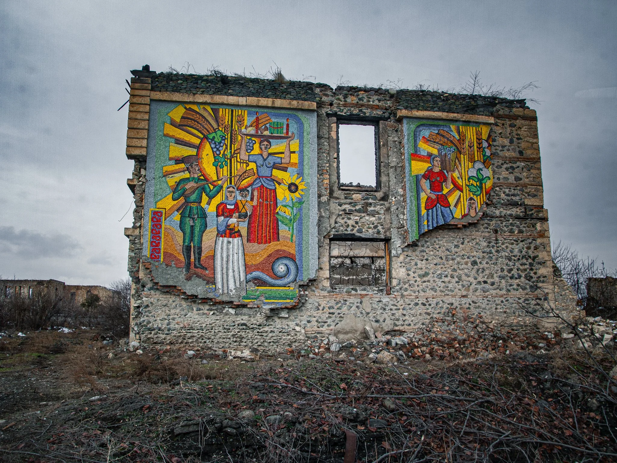 Mural painting on a dilapidated building wall depicting historical or mythological figures with bright colors, set outdoors under a cloudy sky.