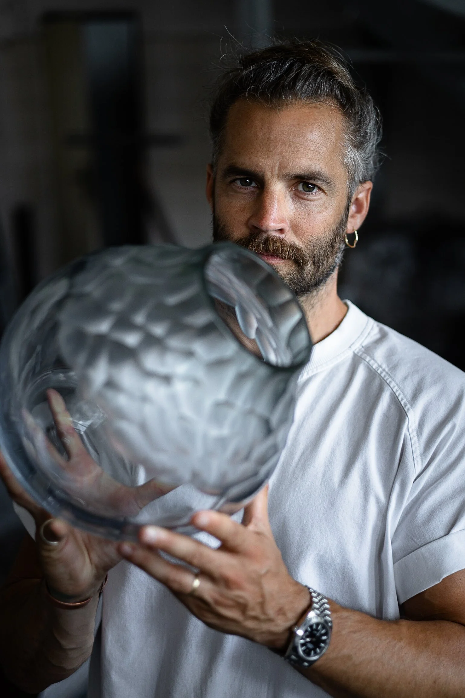 A man with a beard and an earring holding a large glass sculpture. He is wearing a white T-shirt, a wristwatch, and a ring, and looking at the camera.