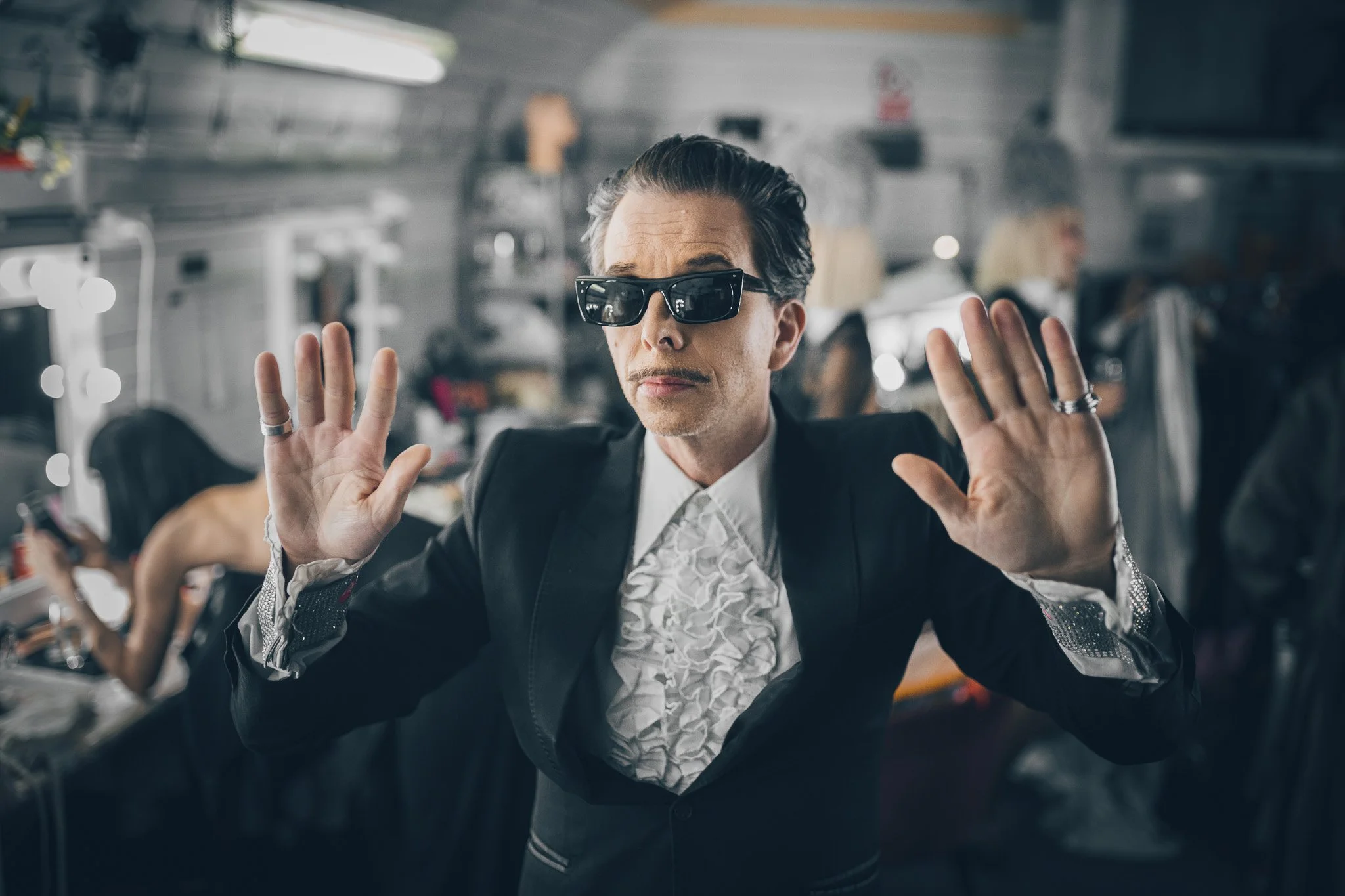 A man wearing dark sunglasses and a black suit with a ruffled white shirt, holding his hands up with palms facing outward in front of a dressing room mirror with bright lights.