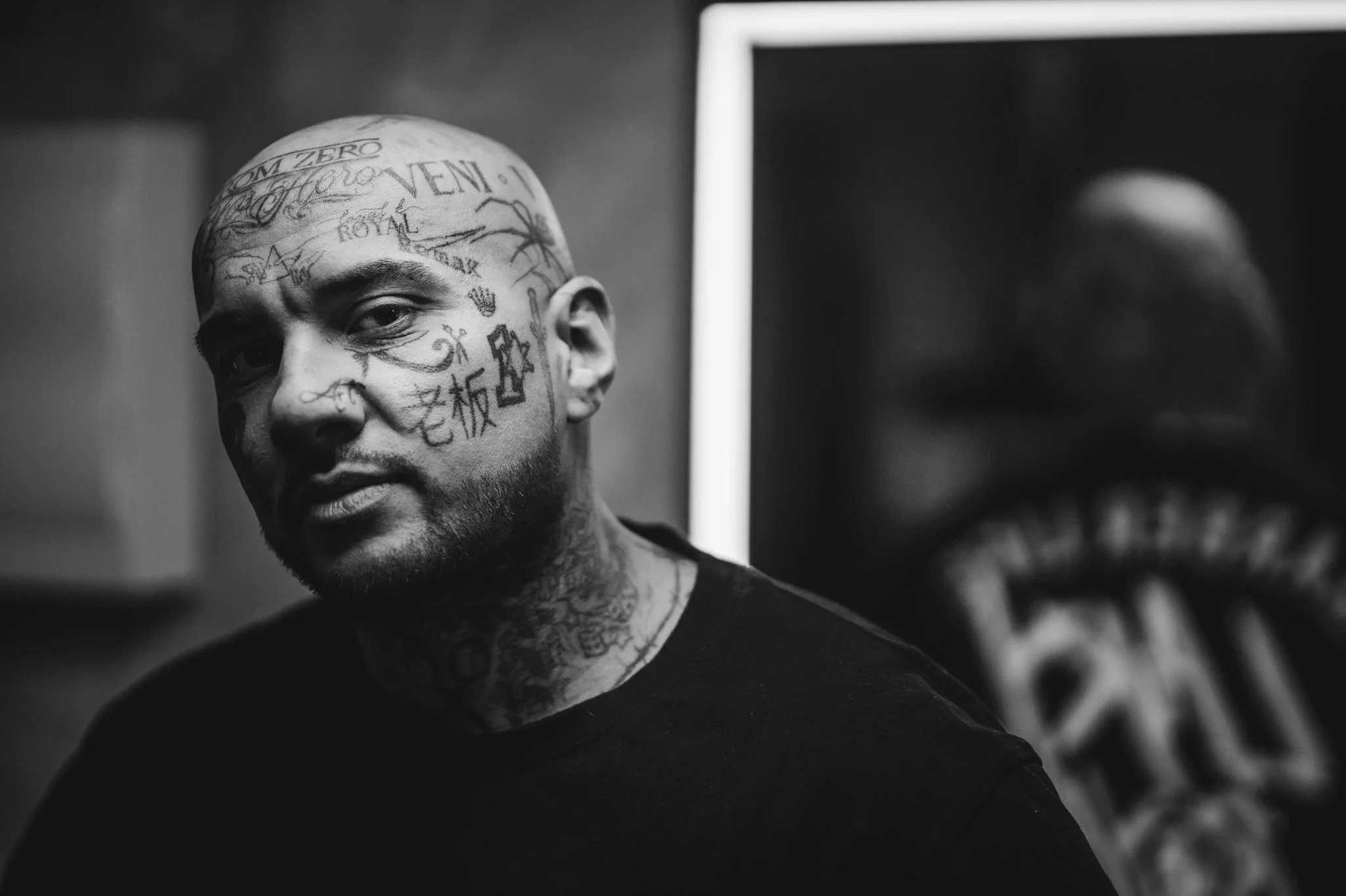 Black and white photograph of a tattooed man with a shaved head, looking at the camera, with tattoos on his face and neck, and a blurred artwork seen in the background.