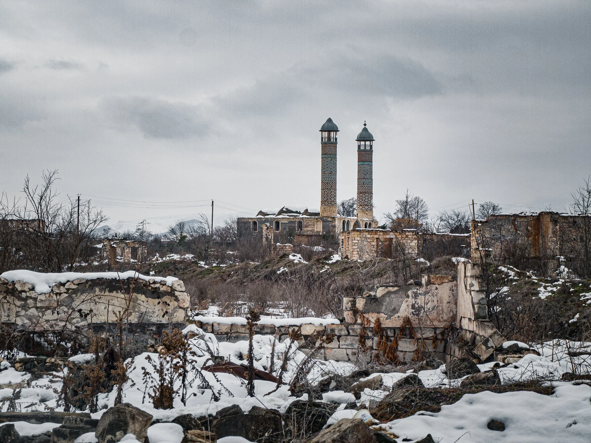 Ruined buildings and leafless trees in a snowy landscape with a structure featuring two decorated chimneys and a cloudy sky in the background.