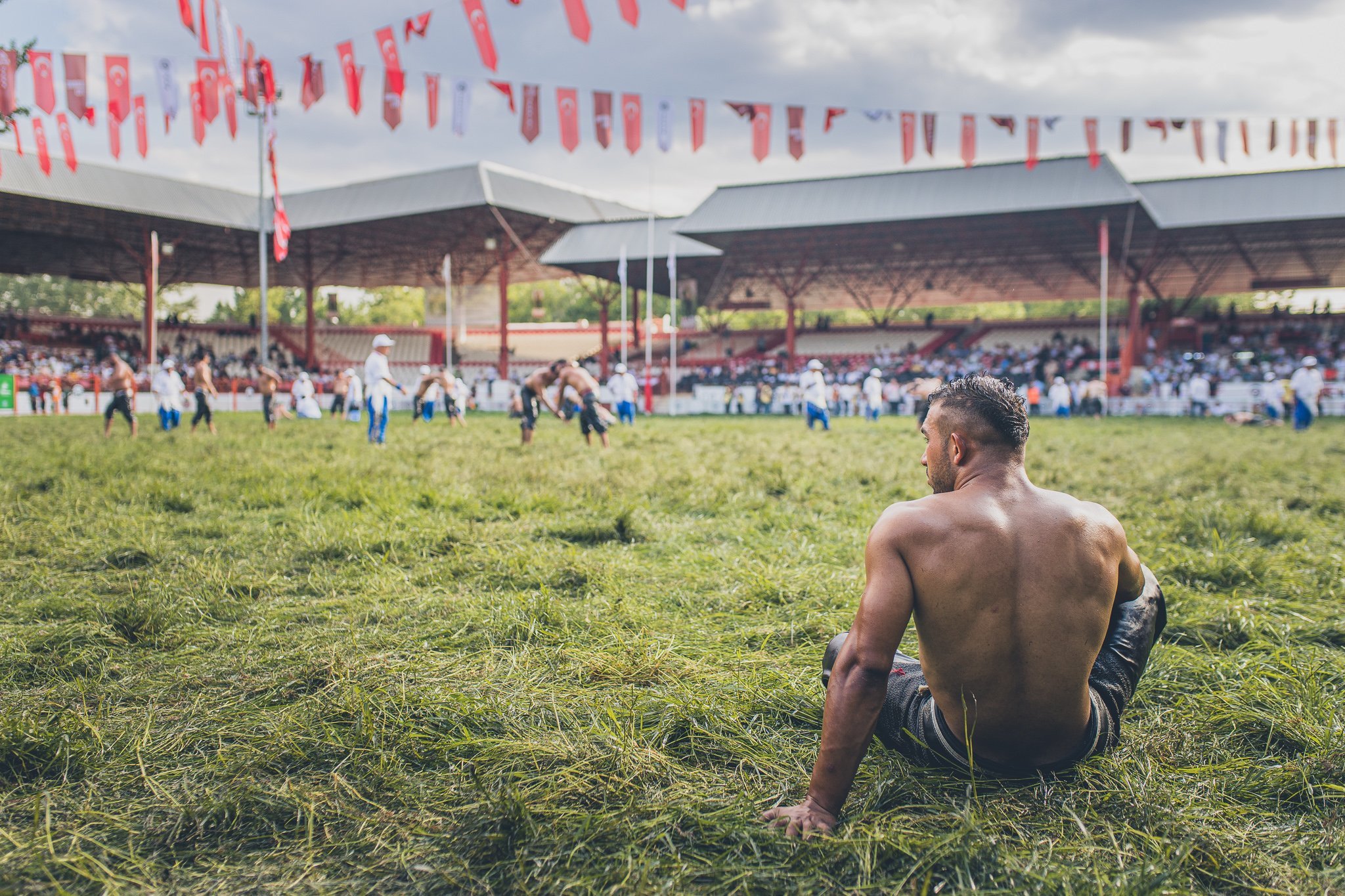 A shirtless man sitting on grass watching a traditional Mexican rodeo event with cowboys in the background