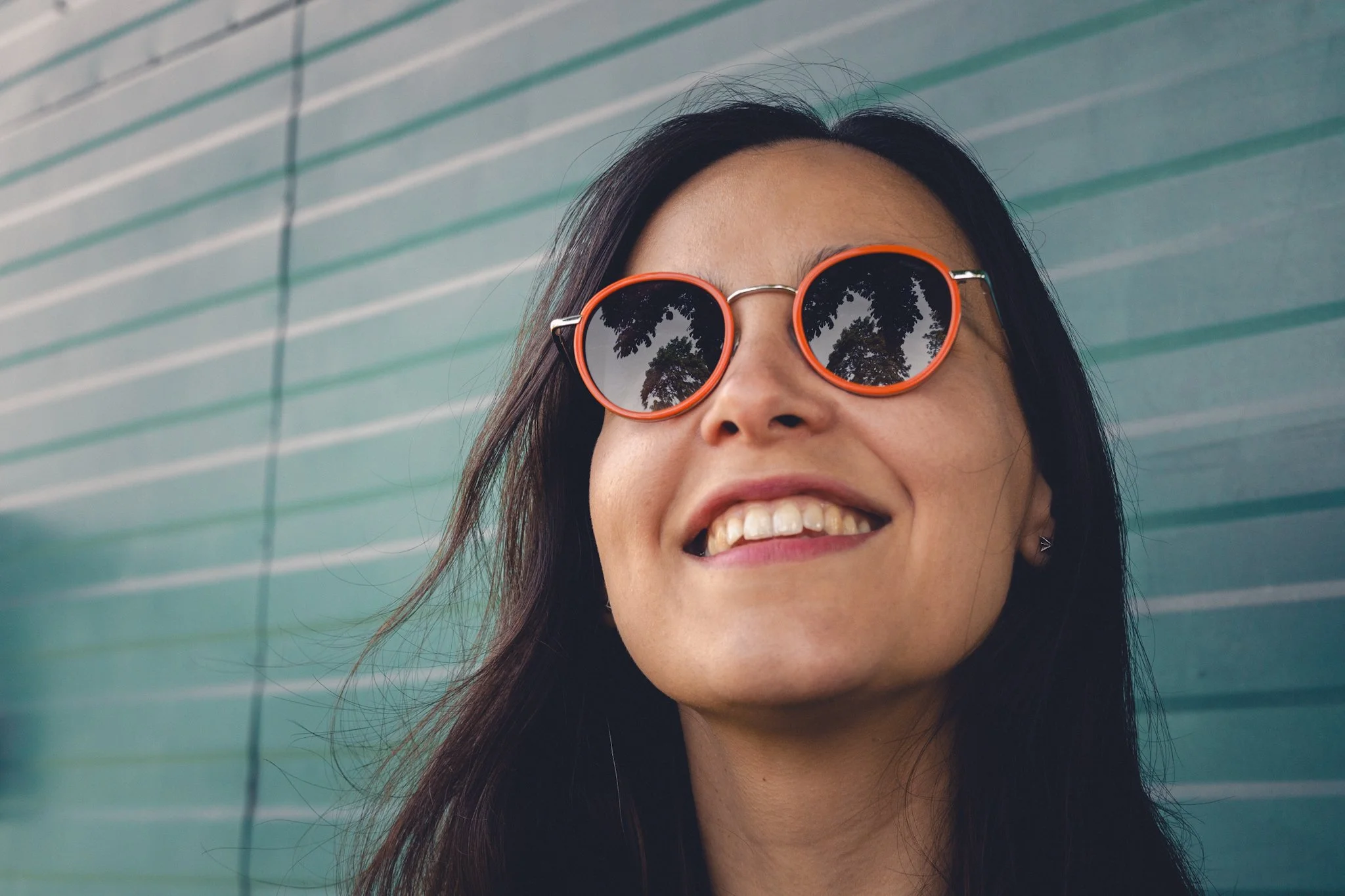 Close-up of a woman smiling, wearing orange sunglasses and standing outdoors against a teal wall with horizontal lines.