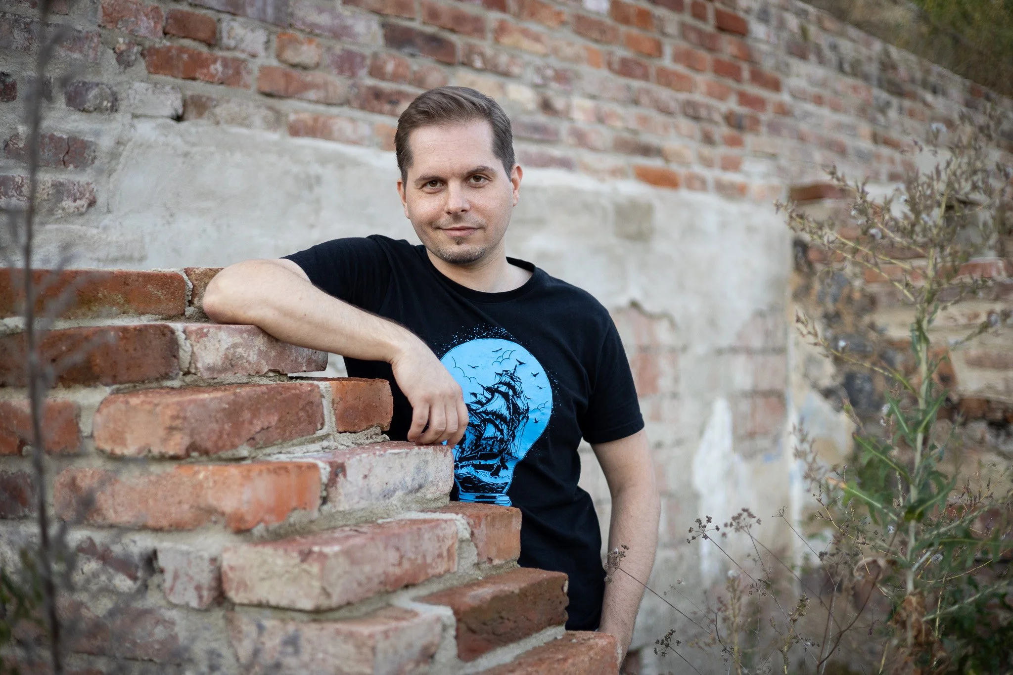 A man in a black t-shirt with a blue graphic of a ship at night, leaning on a brick wall outdoors next to some plants with a brick and concrete wall background.