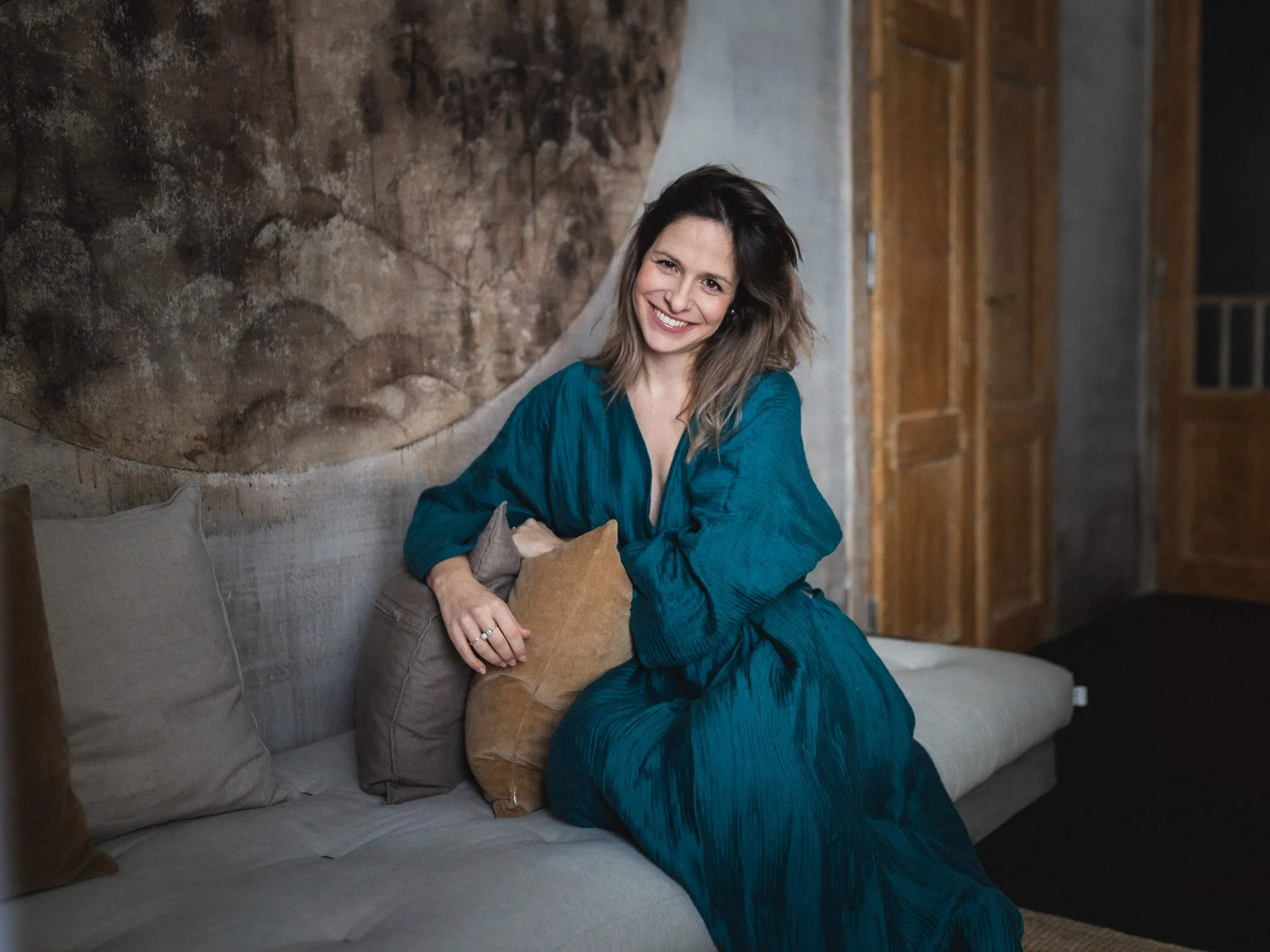 Smiling woman in a blue dress sitting on a sofa with pillows in a rustic room with wooden and stone elements