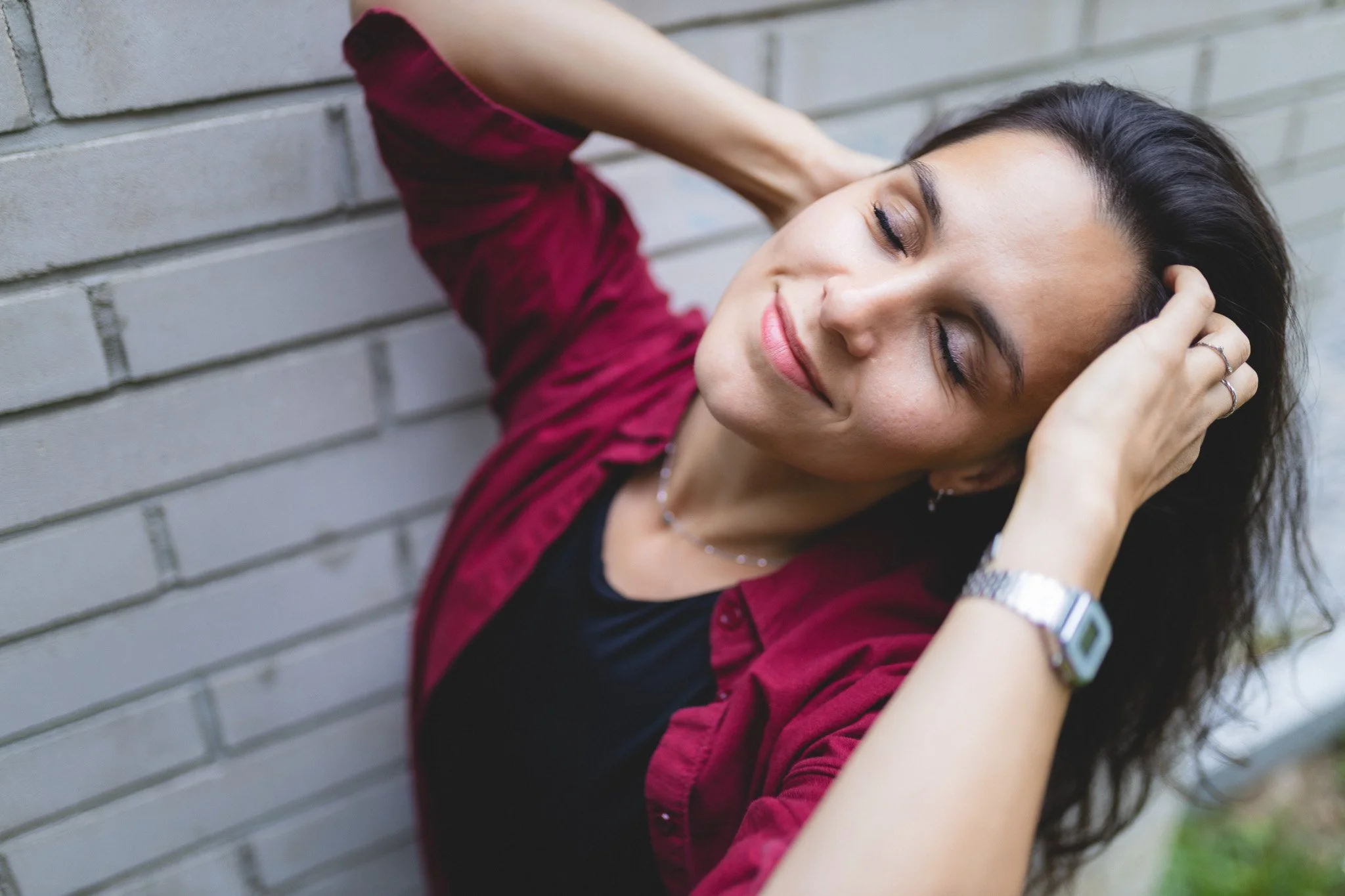 A woman with dark hair and a red shirt smiling with her eyes closed against a gray brick wall.