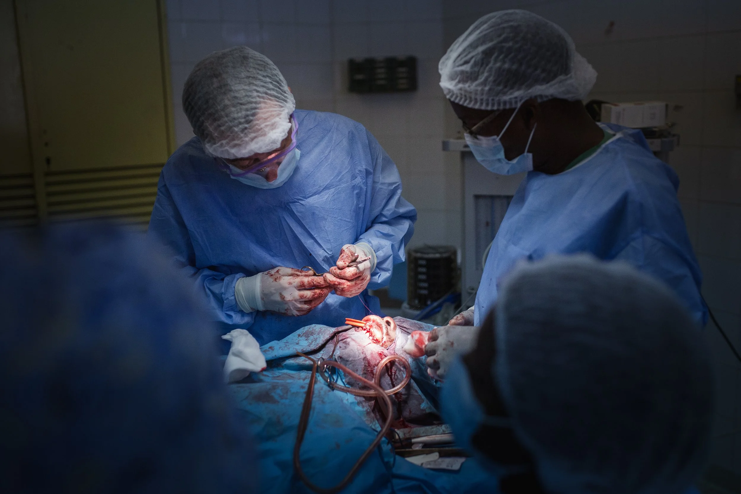 Medical team performing surgery on a patient in an operating room, wearing blue scrubs, masks, and hair coverings.