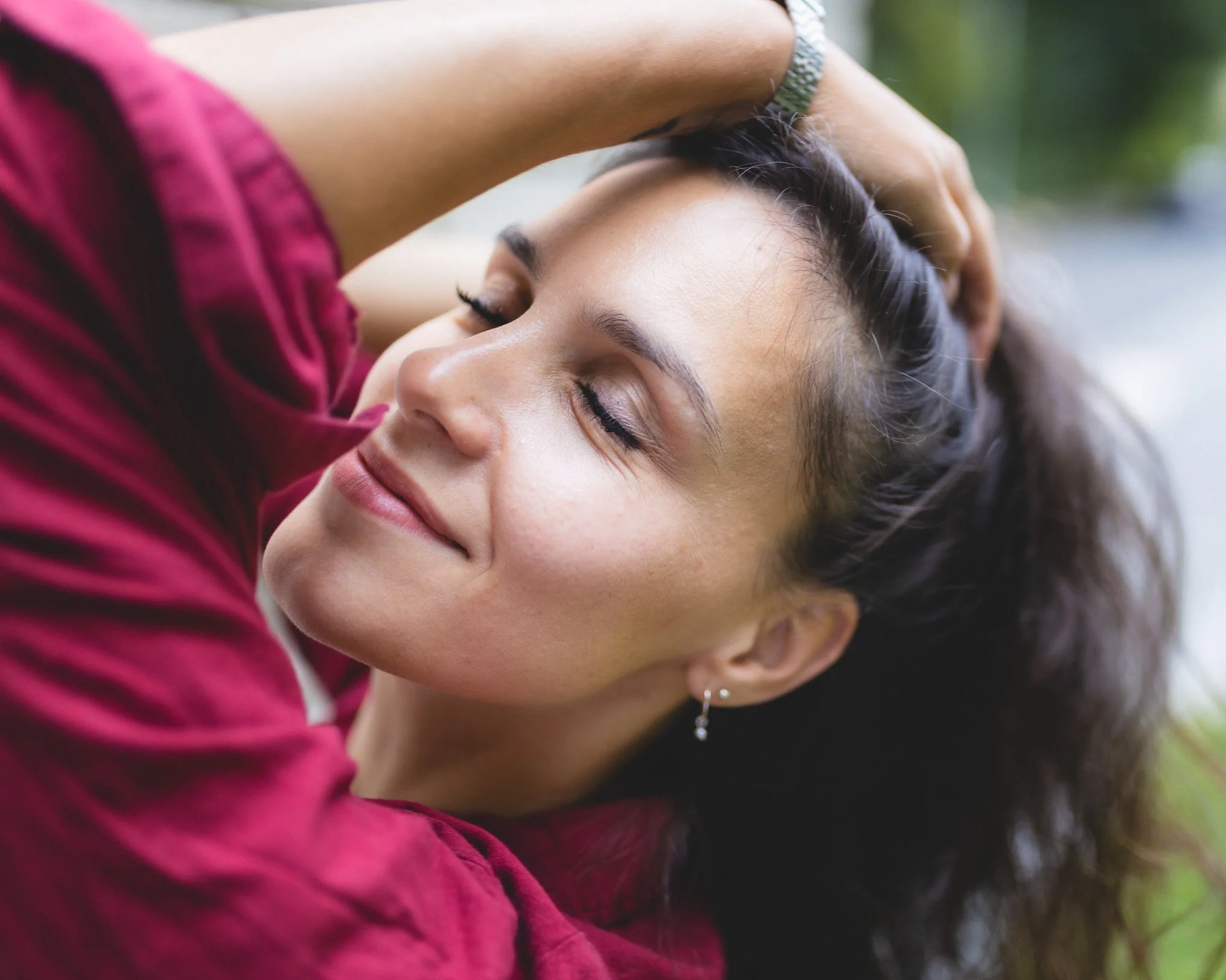Close-up of a woman relaxing outdoors with her eyes closed, smiling gently, wearing a pink top, with her hand in her hair.