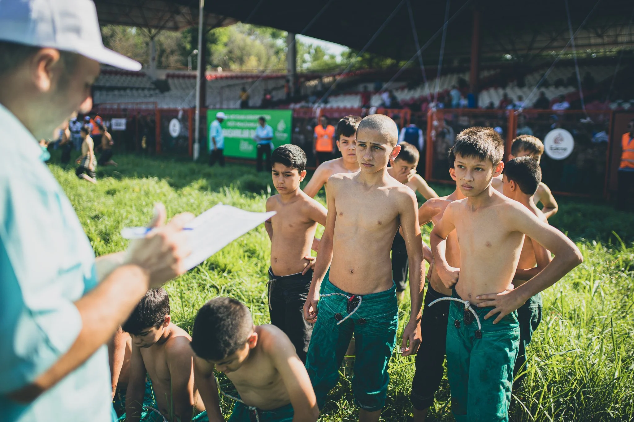 Group of young boys shirtless, standing on grass, listening to a person with a clipboard at an outdoor event or competition.