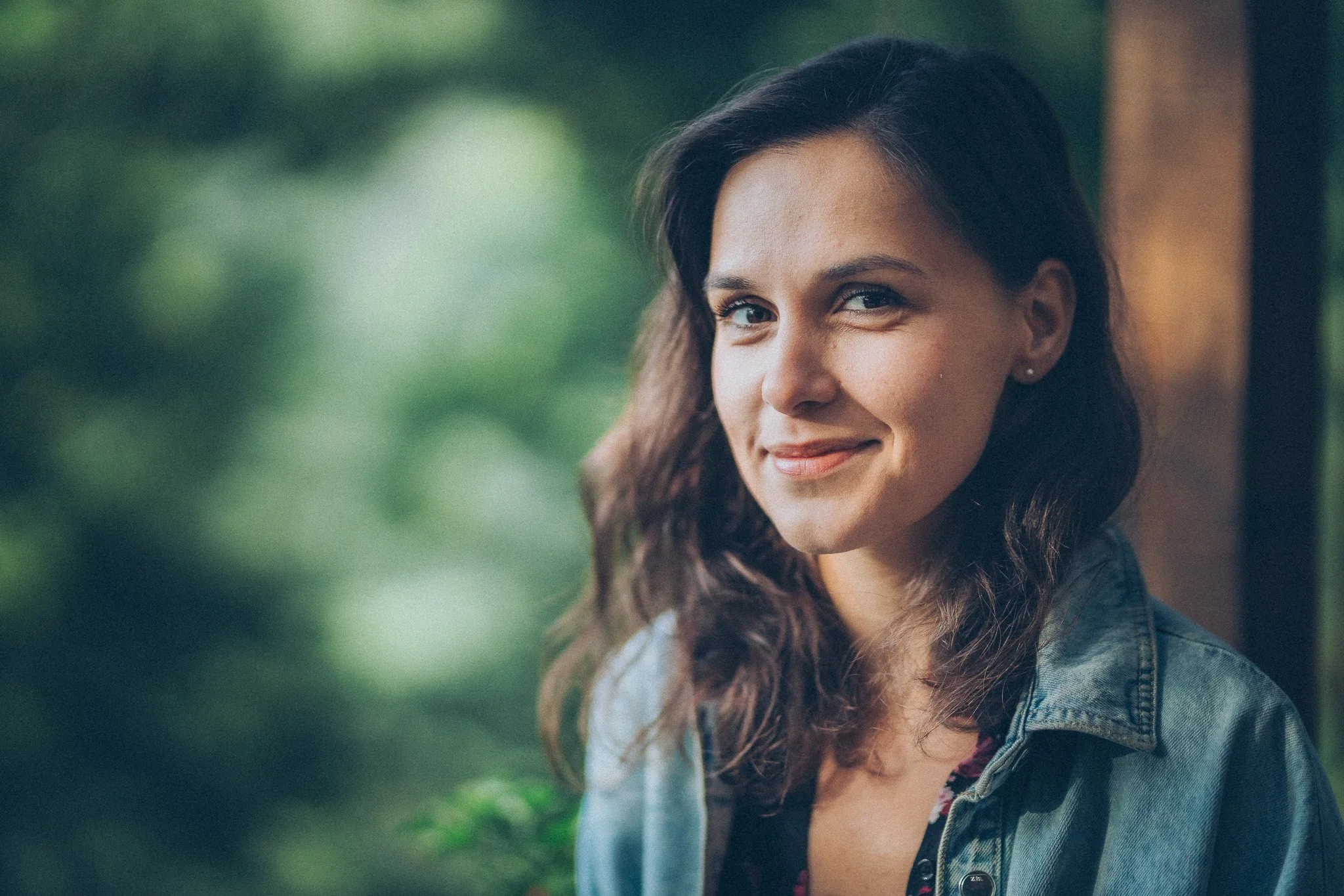 A woman with wavy dark hair, wearing a denim jacket, smiles softly outdoors with greenery in the background.