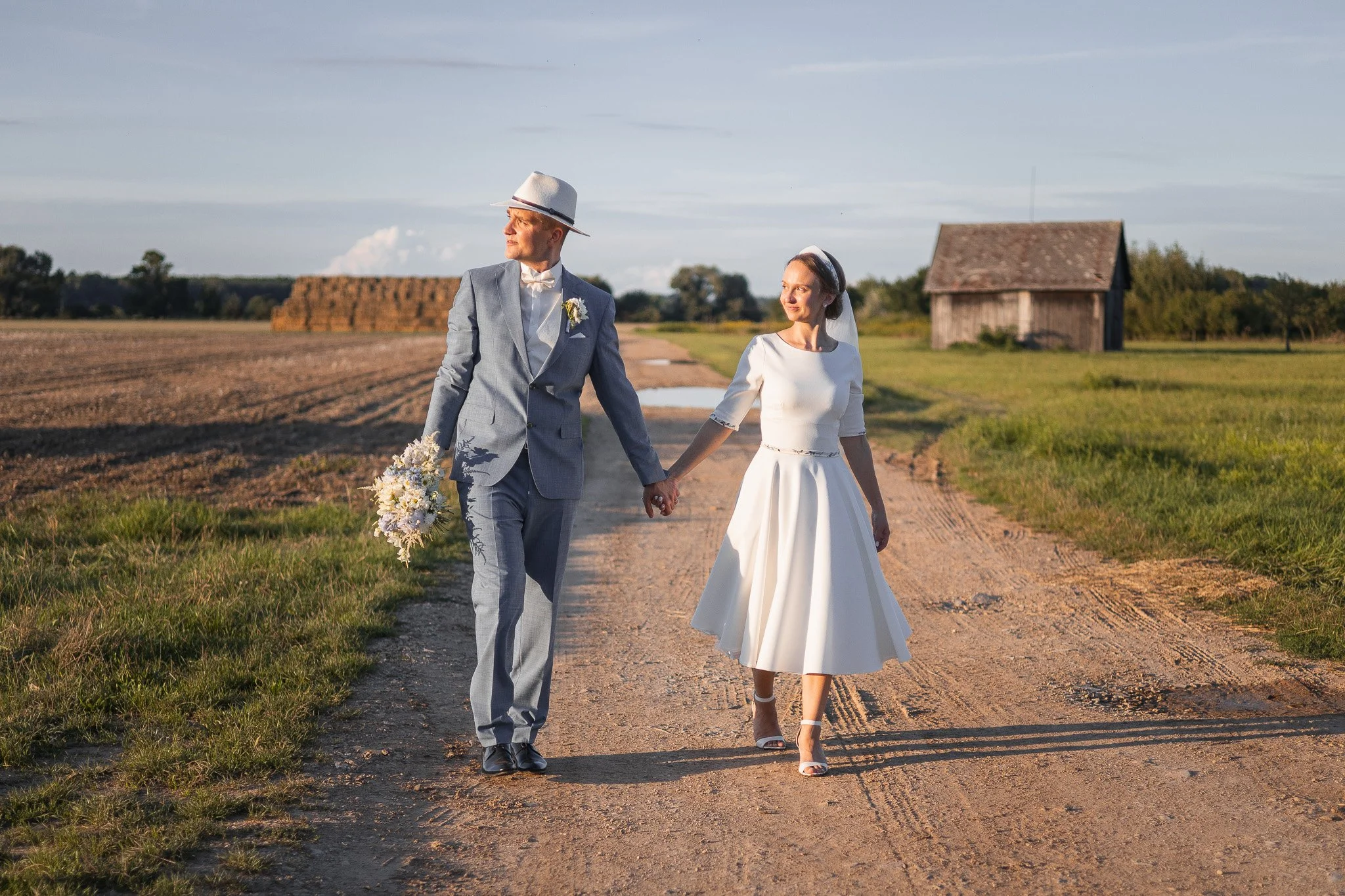A bride and groom walking hand in hand on a rural dirt road with a barn and hay in the background during sunset.