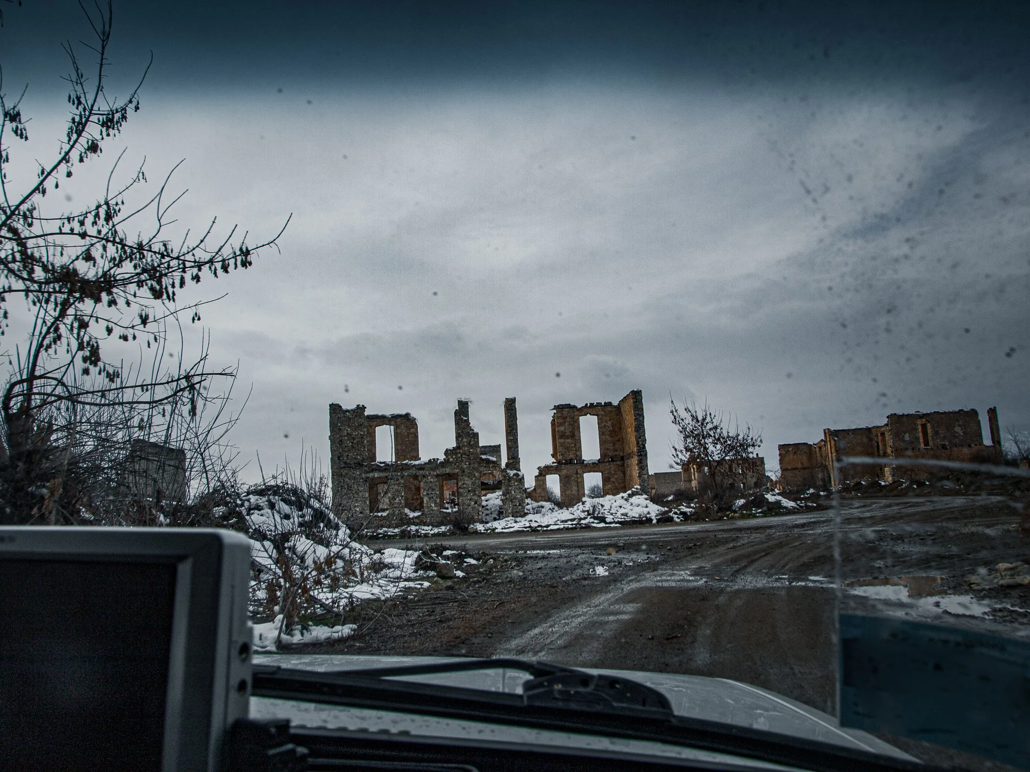 Ruins of an old stone building on a snowy landscape, seen from inside a vehicle, with overcast sky and leafless trees.
