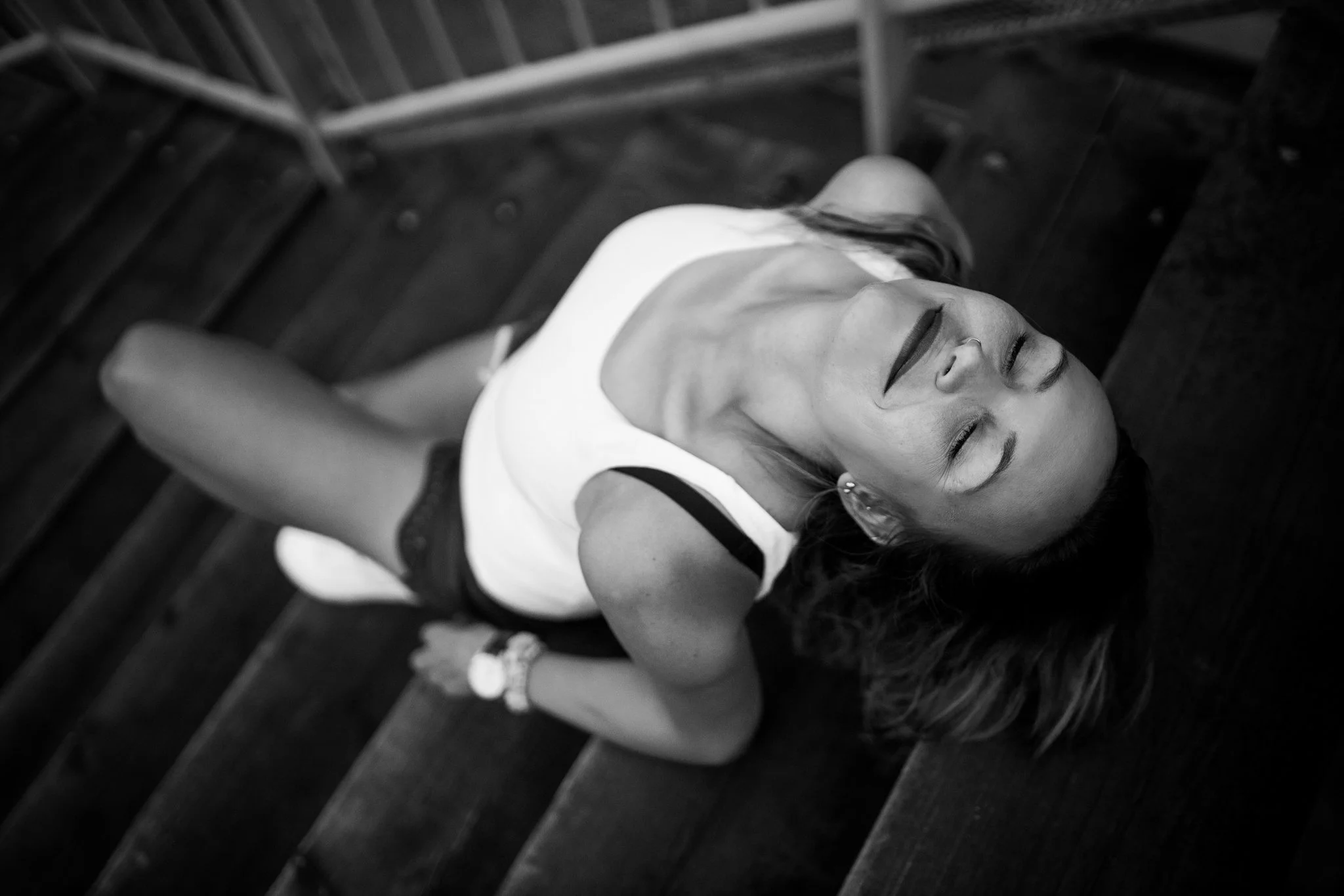 Black and white photo of a woman lying on her back with eyes closed, smiling, on wooden stairs, wearing a sleeveless white top and dark shorts, with her hair spread around her head.