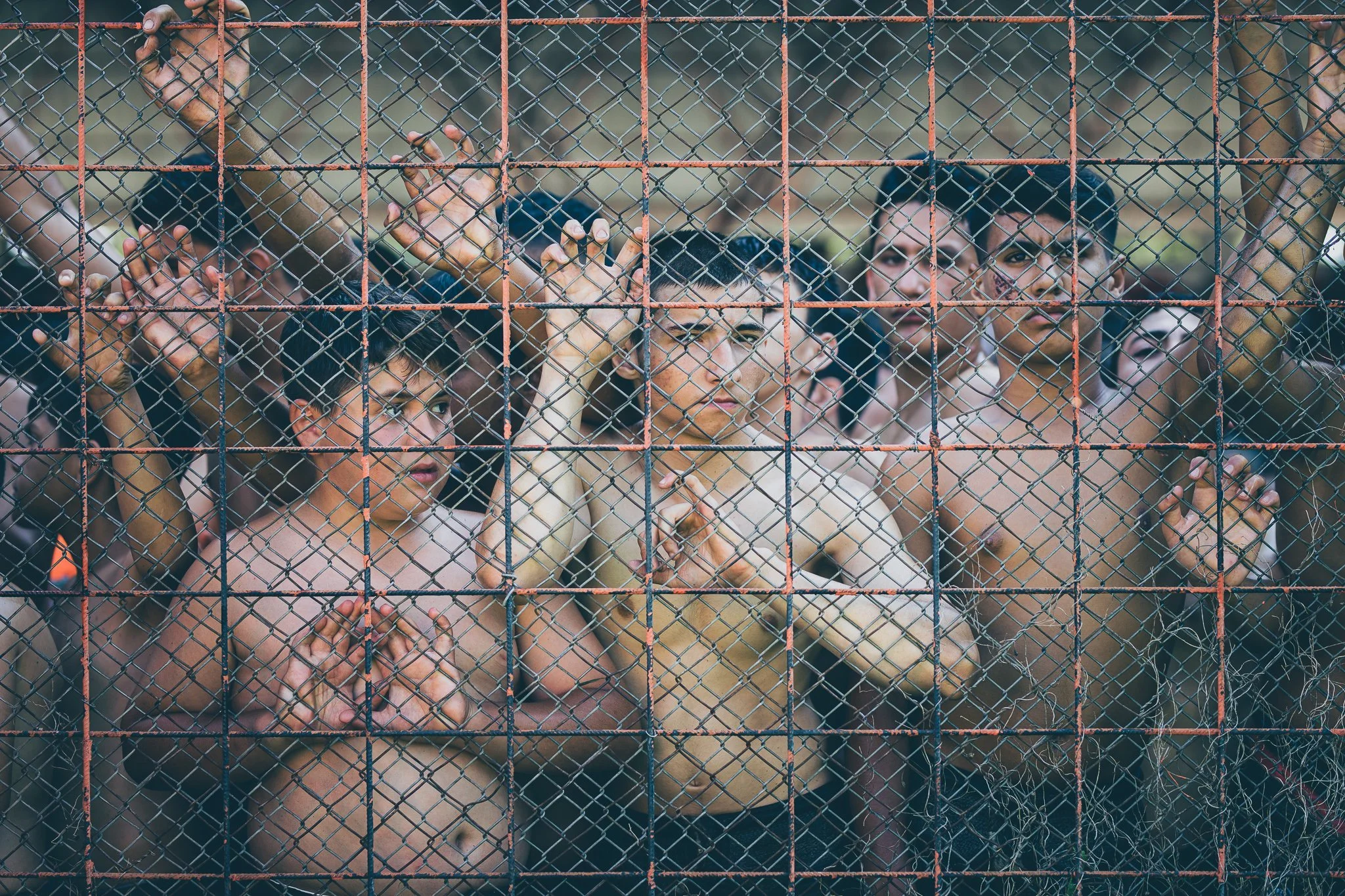 A group of shirtless men behind a rusted chain-link fence, holding onto the fence with their hands, and looking out.