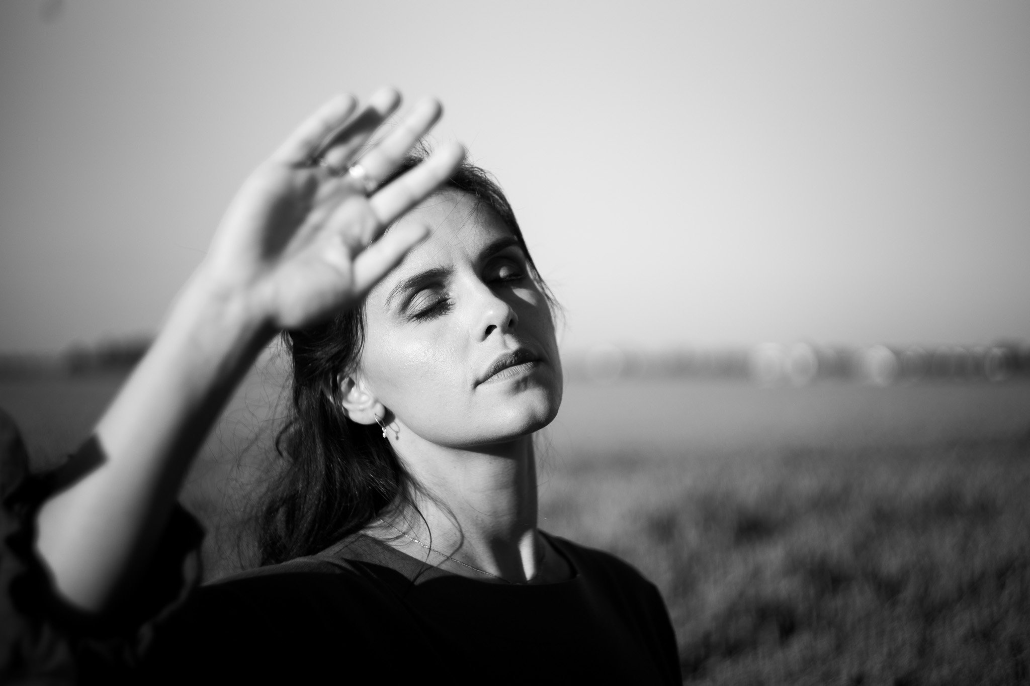 A black and white image of a woman with closed eyes, holding her hand up to her forehead as if shielding her eyes from the sun, in an outdoor setting with fields in the background.