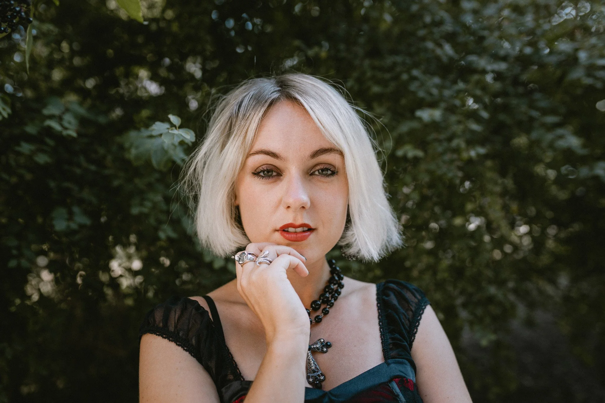 A woman with short blonde hair and blue eyes standing outdoors in front of green foliage, wearing black lace top and jewelry.