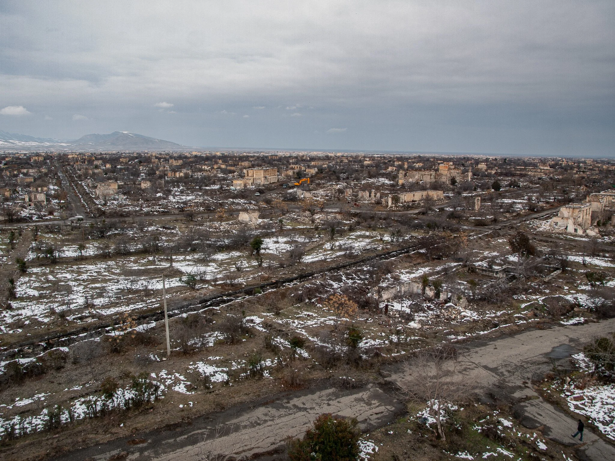 A wide view of a city landscape in winter with snow patches, broken buildings, and overcast sky, with distant mountains.