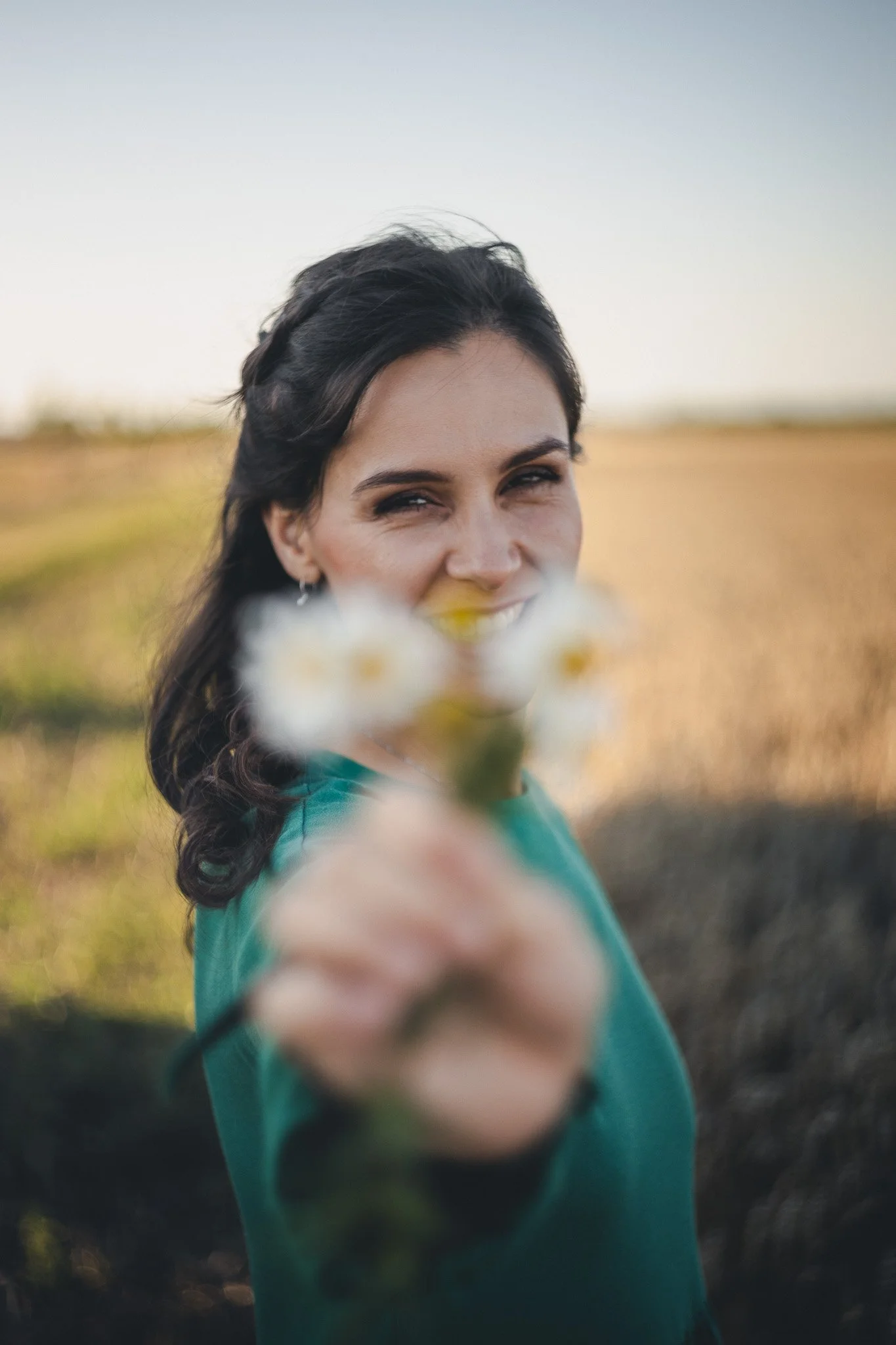 A woman with dark hair smiling and holding a small bouquet of white flowers towards the camera in an outdoor setting during daytime.