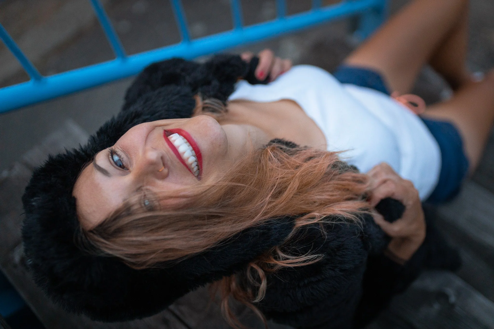 A woman with colored hair laying on her back on wooden planks, smiling toward the camera, wearing a white top and dark shorts, holding a black dog close to her face, with a blue railing in the background.