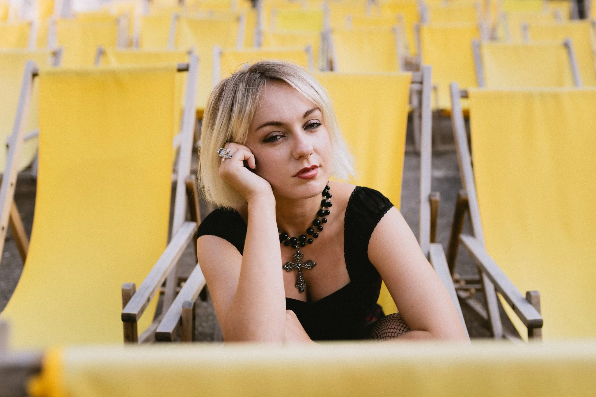 Young woman with blonde hair sitting among yellow chairs, wearing black clothing, a large cross necklace, and fishnet stockings, resting her head on her hand with a pensive expression.