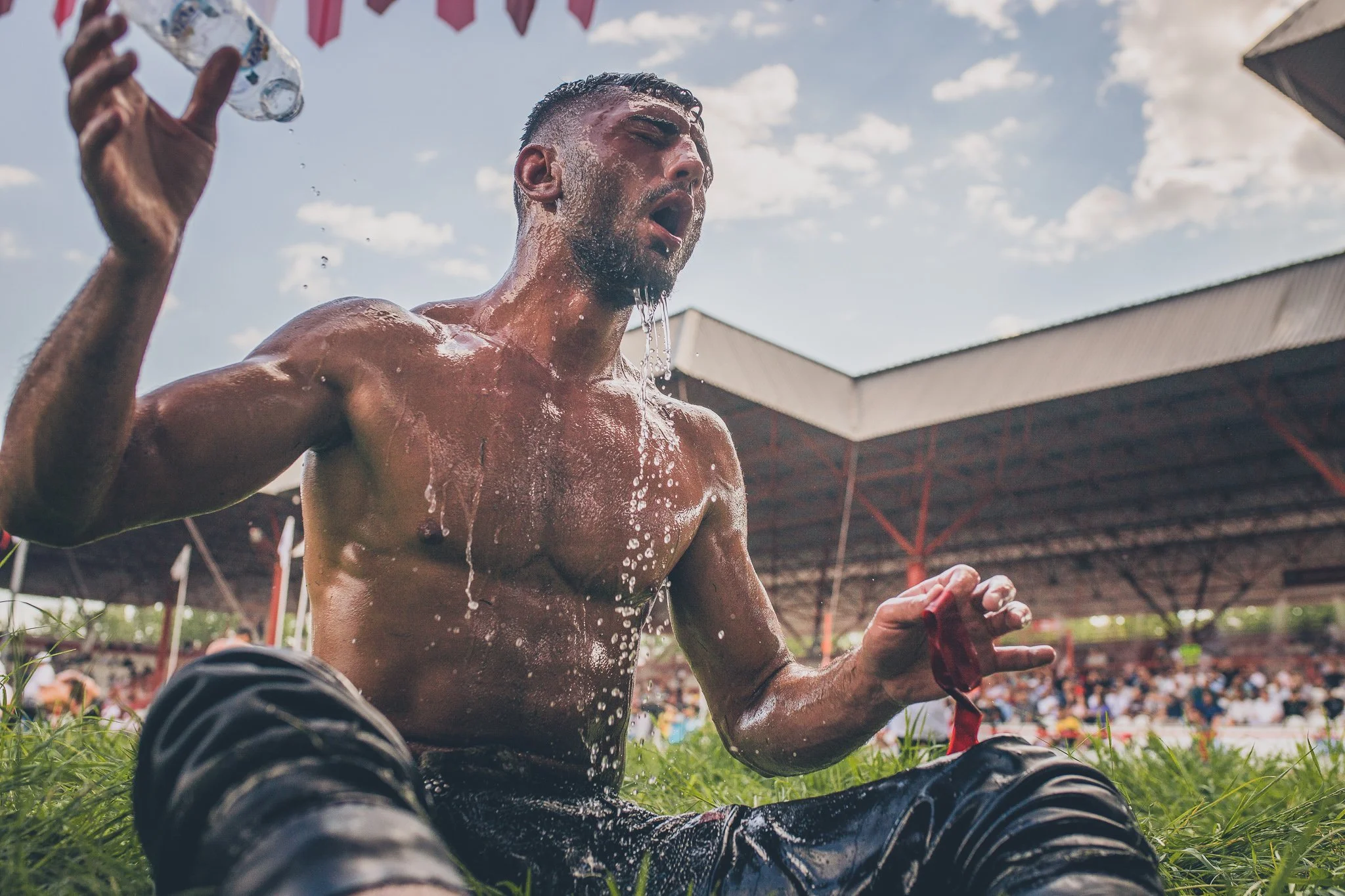 A shirtless man sitting on the grass, drinking water from a plastic bottle after a sports event, with a crowd of people and a stadium roof in the background.