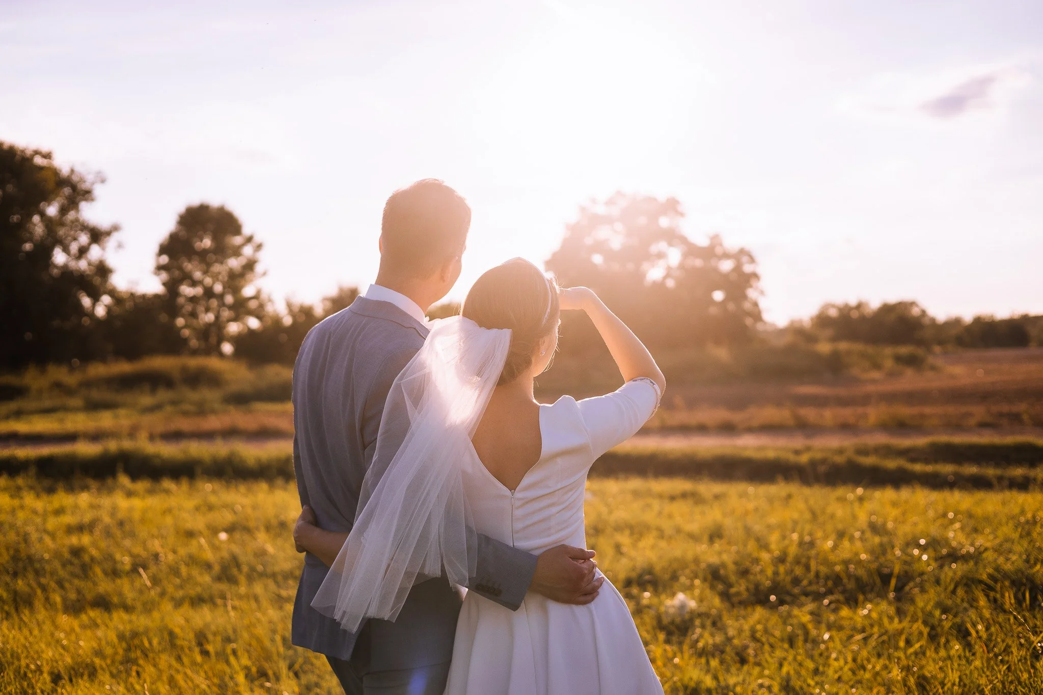 A bride and groom standing outdoors in a field at sunset, sharing a moment together.