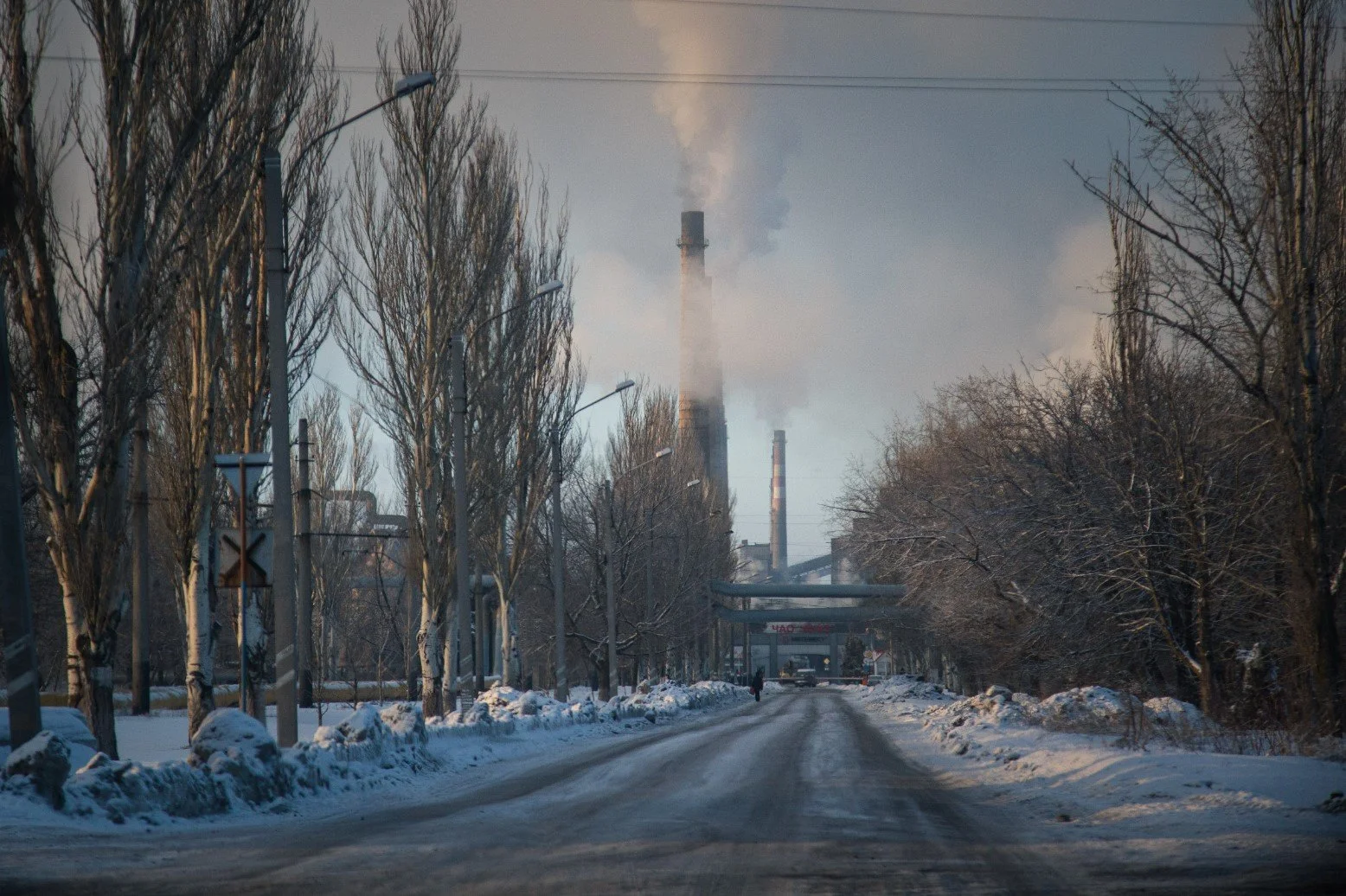 A snow-covered road flanked by leafless trees, with industrial chimneys emitting smoke in the background during winter.