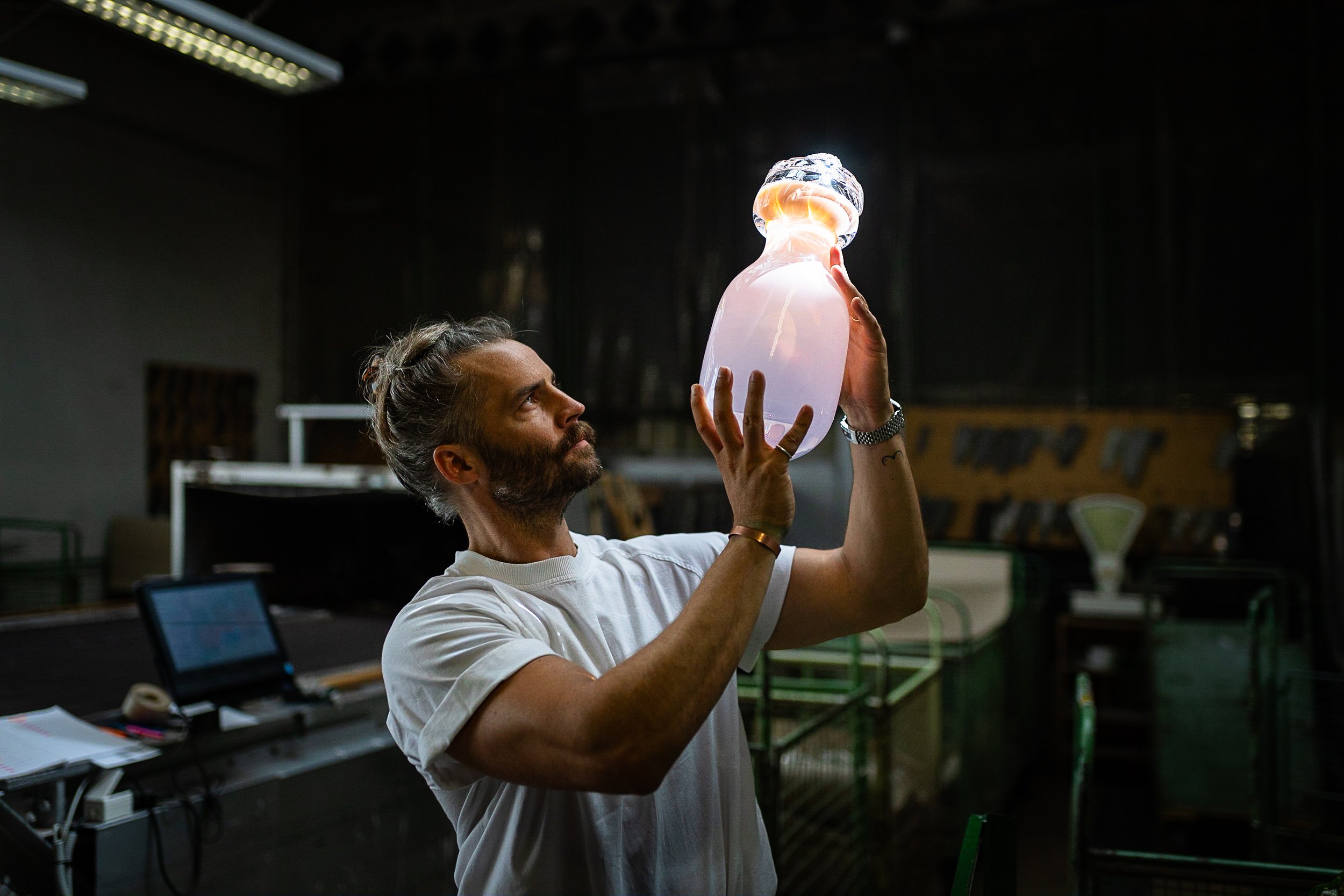 A man with long hair and a beard holding a glowing, translucent glass perfume bottle indoors with industrial lighting.