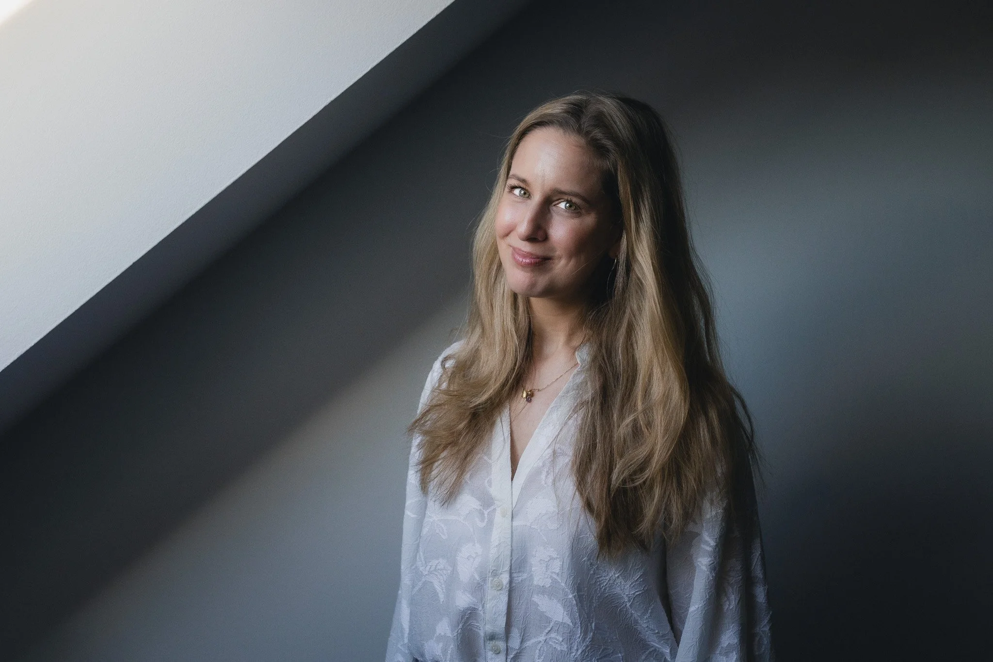 A woman with long, wavy blonde hair wearing a white button-up shirt standing by a window with sunlight streaming in.