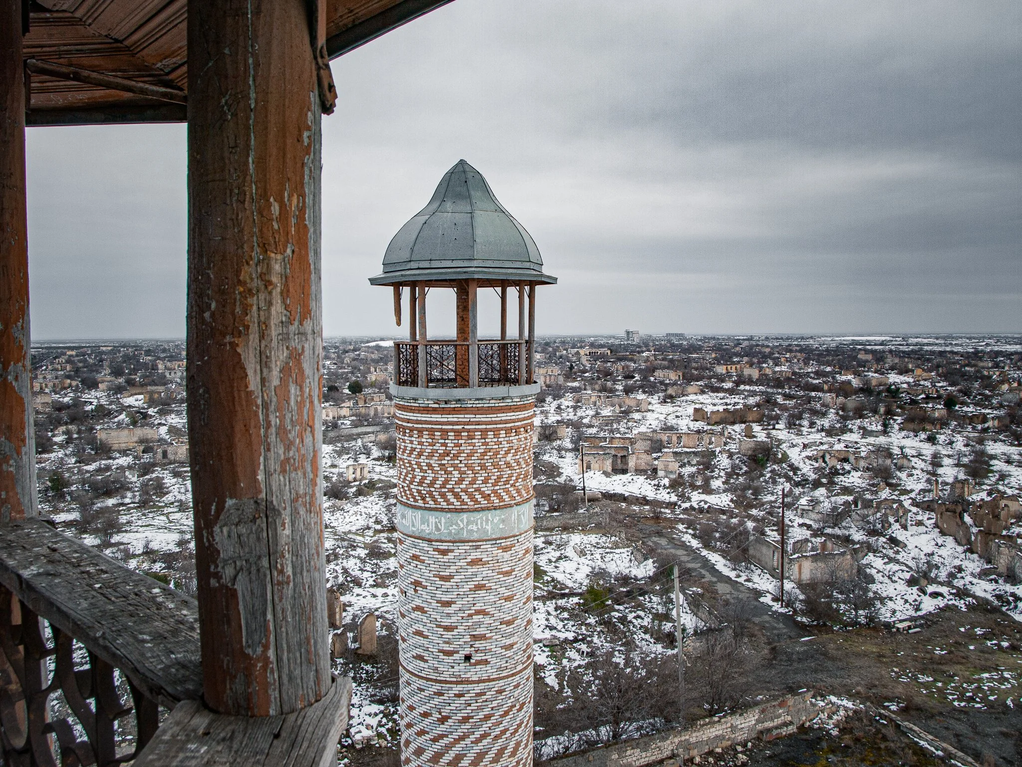 A tall brick minaret with white Arabic script and decorative patterns, topped with a metal dome, overlooking a snow-covered cityscape with damaged buildings under cloudy skies.