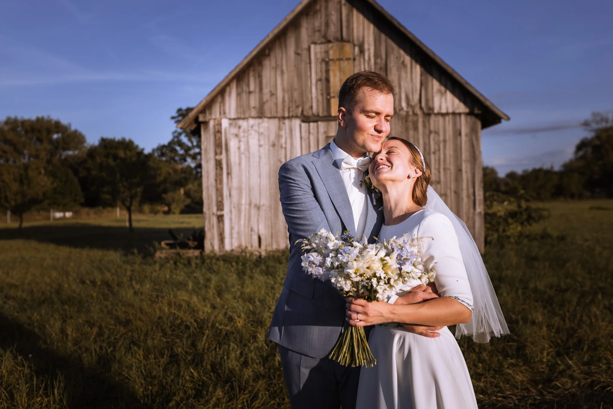 A bride and groom embracing outdoors in front of a rustic wooden barn during sunset, with the bride holding a bouquet of white flowers.