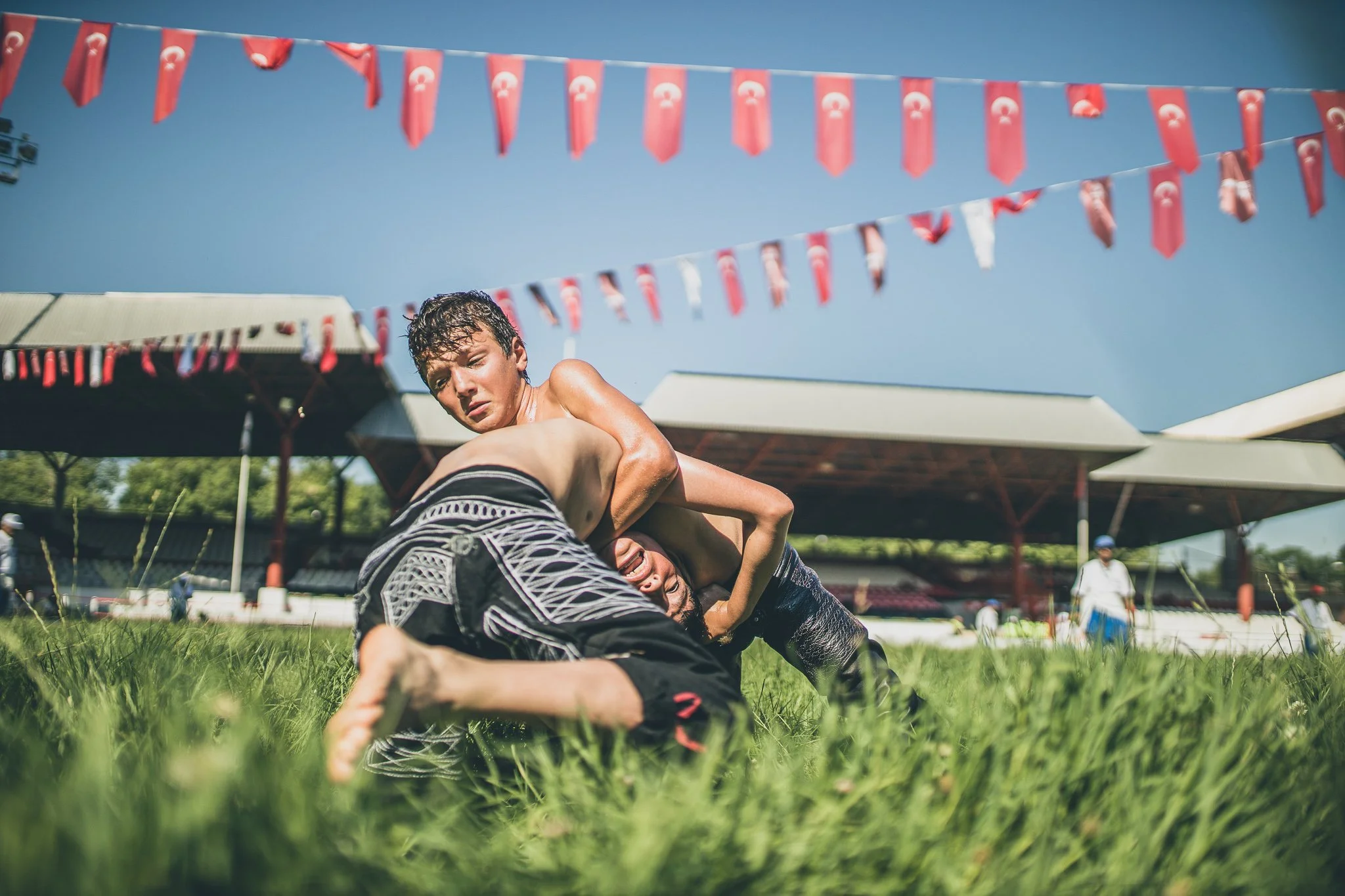 Two shirtless young men wrestling on grass field during outdoor event under blue sky, with red pennant banners and a pavilion in the background.
