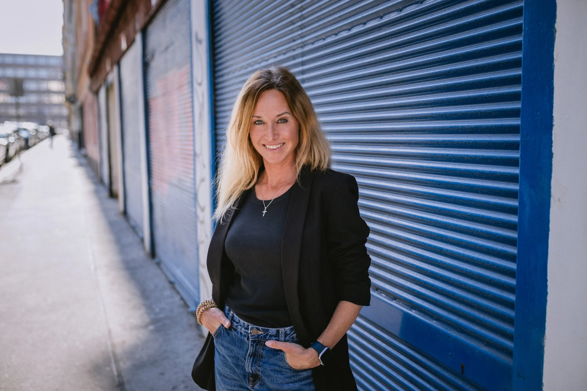 A woman with blonde hair smiling and standing with hands in pockets in front of blue metal shutters on a city sidewalk.