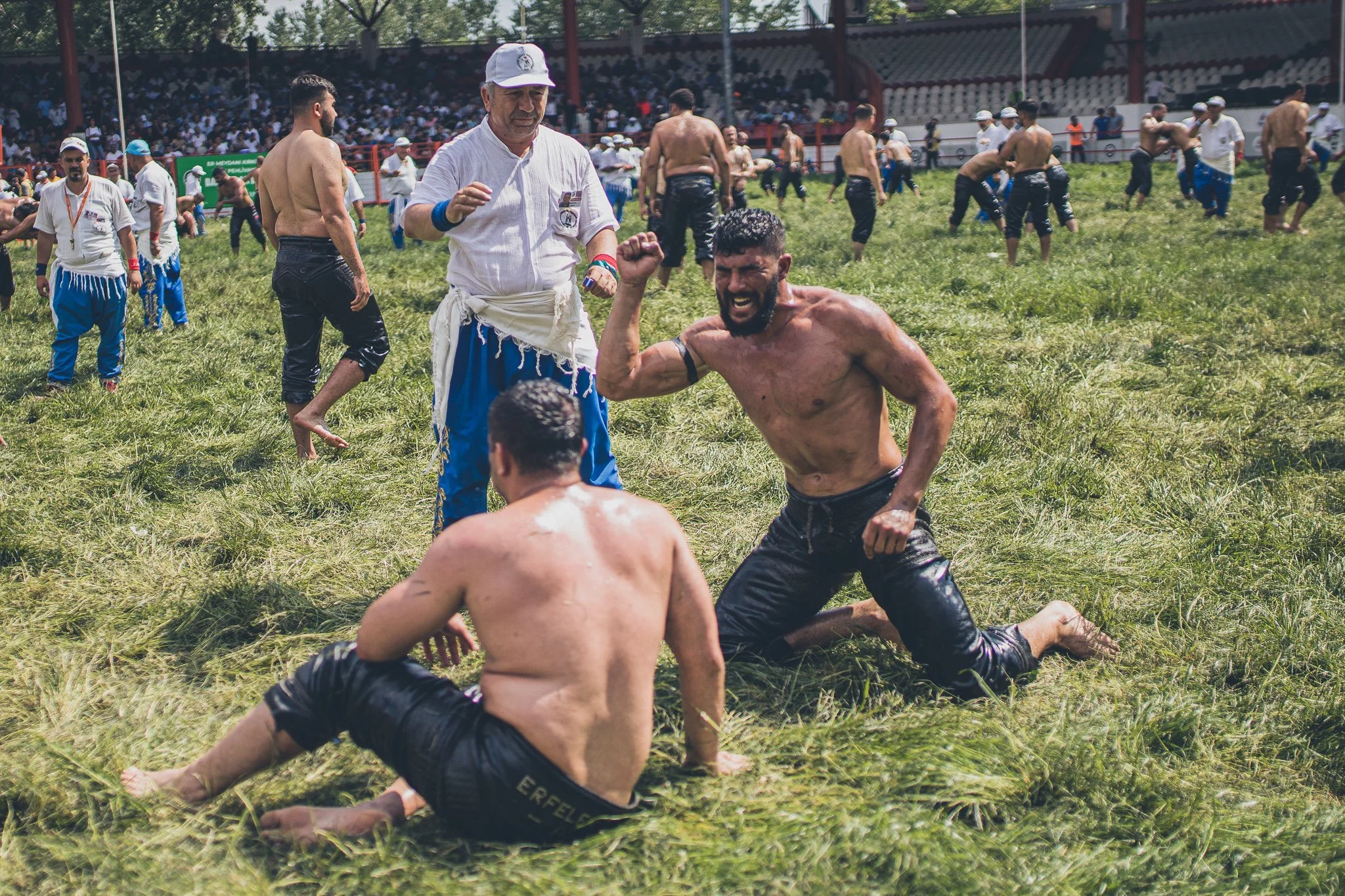 Two shirtless men wrestling on a grassy field while a man in a white shirt and cap watches. People in athletic attire are in the background.