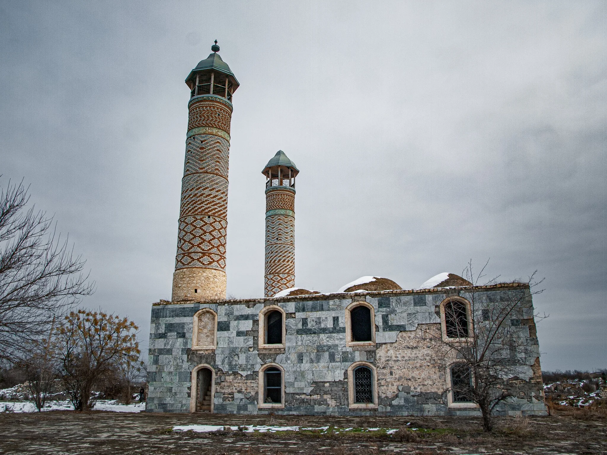 An old stone building with two tall, narrow tower structures featuring colorful, patterned tile designs, set against a cloudy sky with snow on the ground.