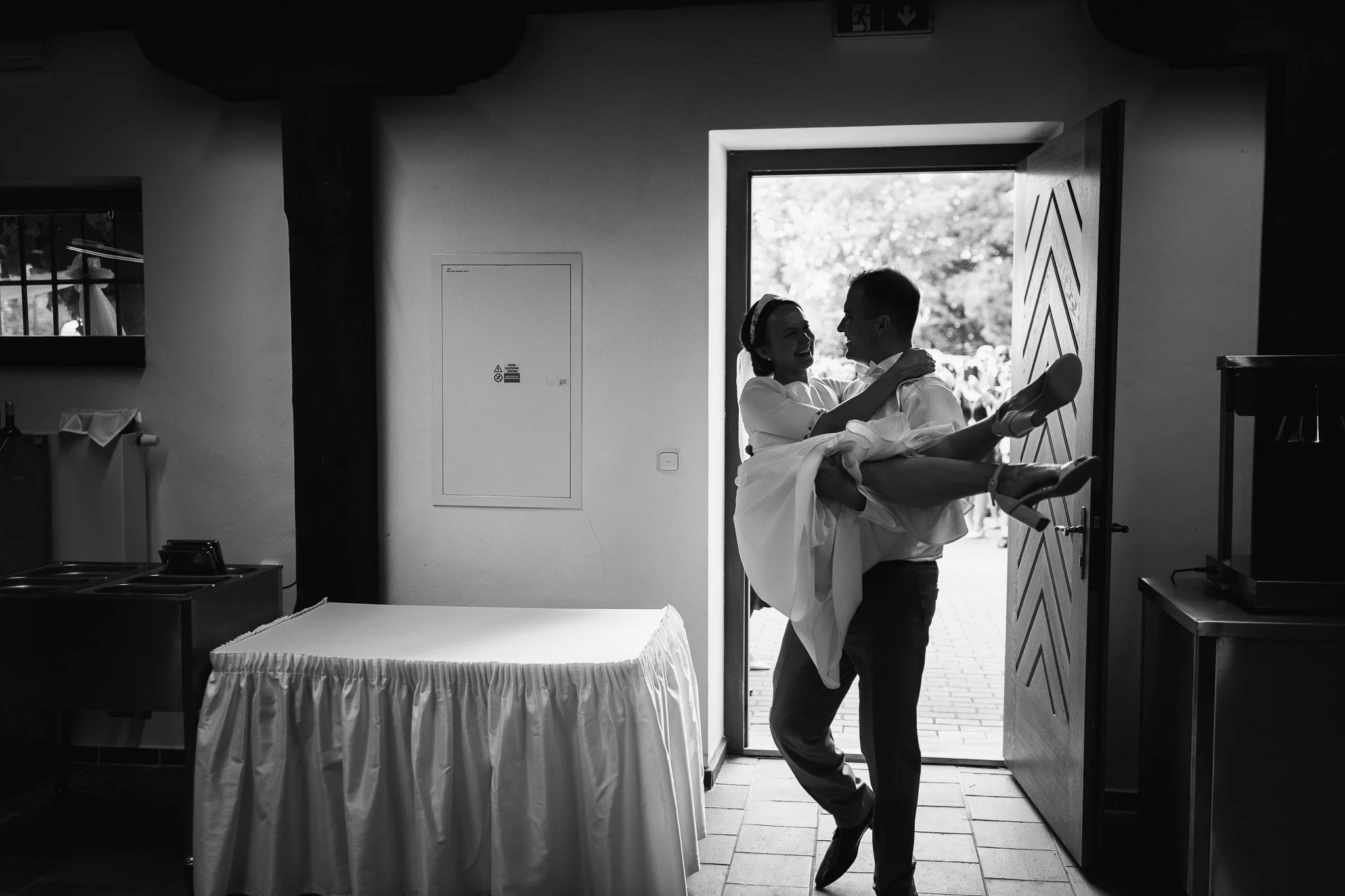 A man carrying a woman in a white dress through a doorway, smiling and embracing, with a group of people outside visible in the background.