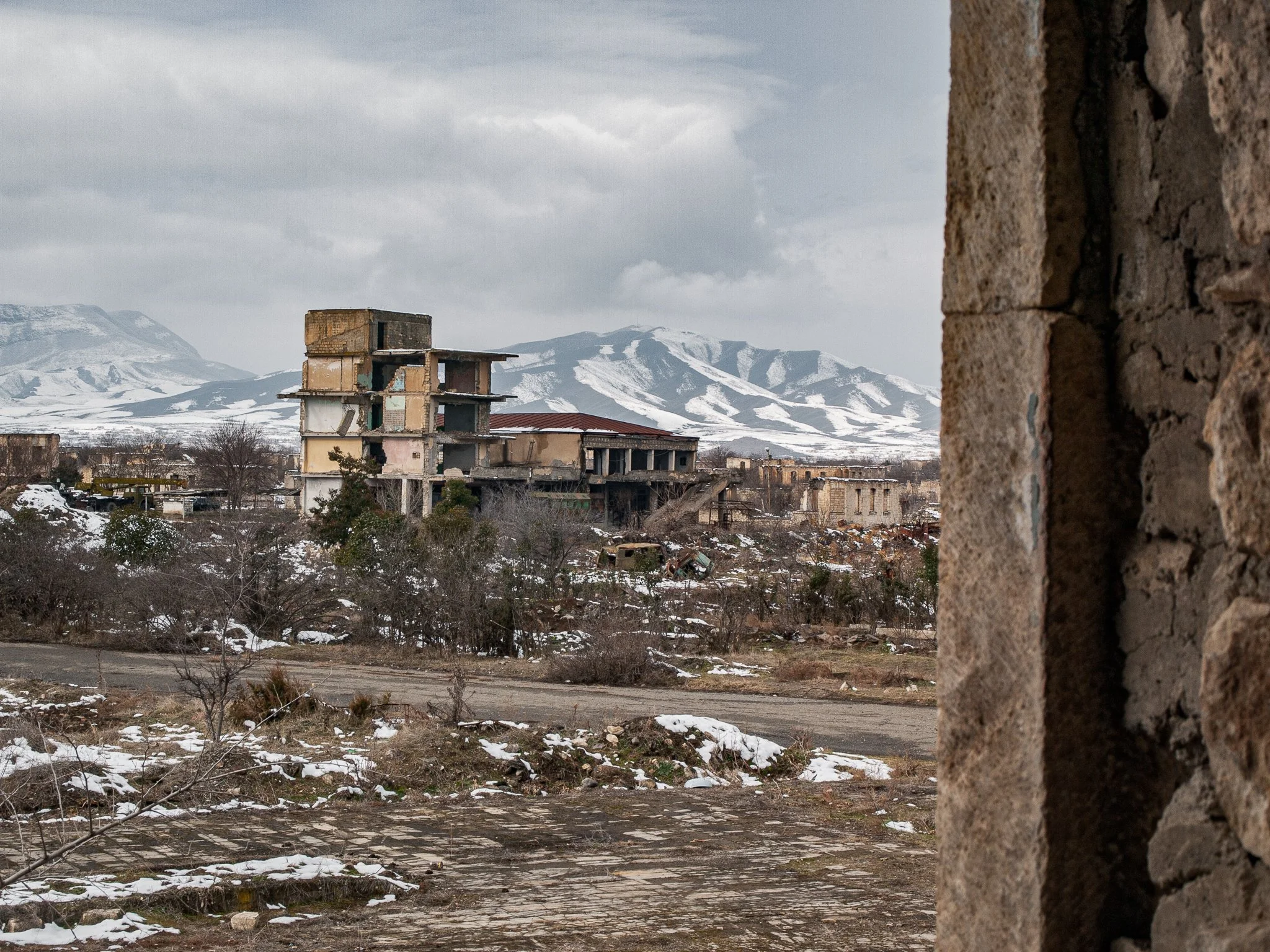 Abandoned multi-story building with broken windows in a snow-covered landscape, with mountains in the background, seen through a window or opening in a stone wall.