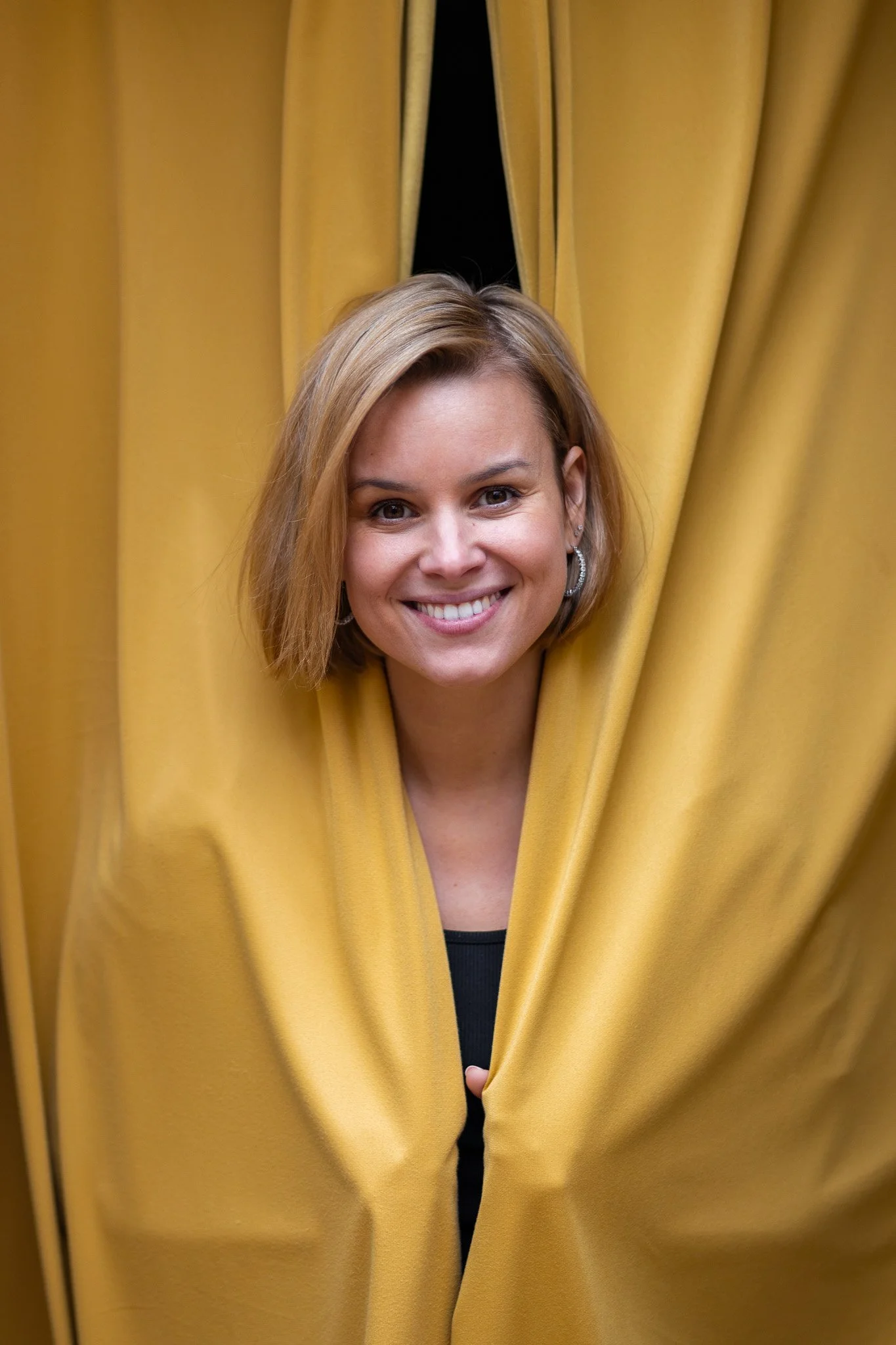 A woman with short blonde hair smiling while peeking through a gap in yellow curtains.